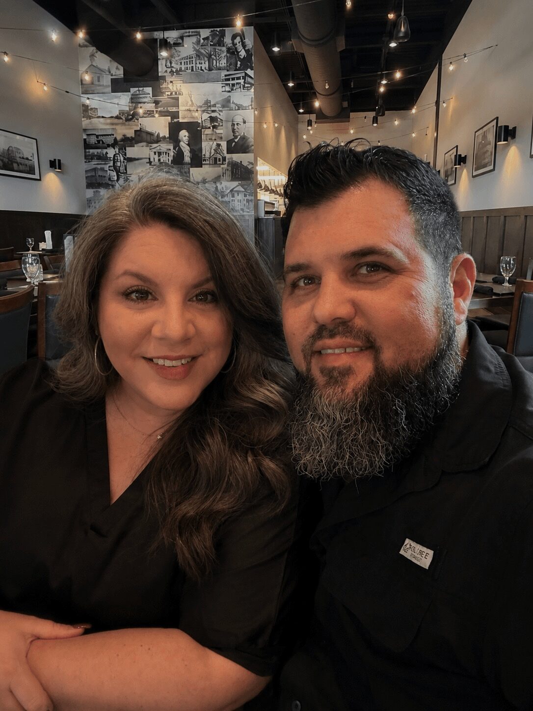 A woman and a man pose together in a restaurant with black walls and photos on the wall behind them.