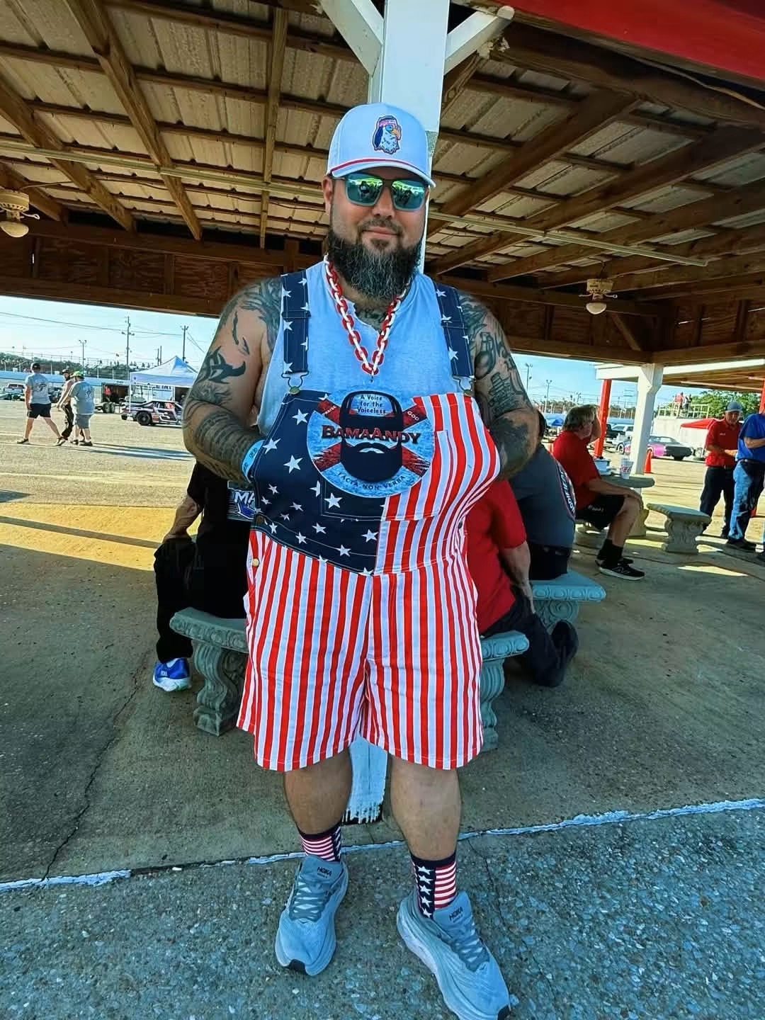 Man with tattoos wearing sunglasses, a cap, and patriotic outfit, standing outdoors under a shelter with people and cars in background.