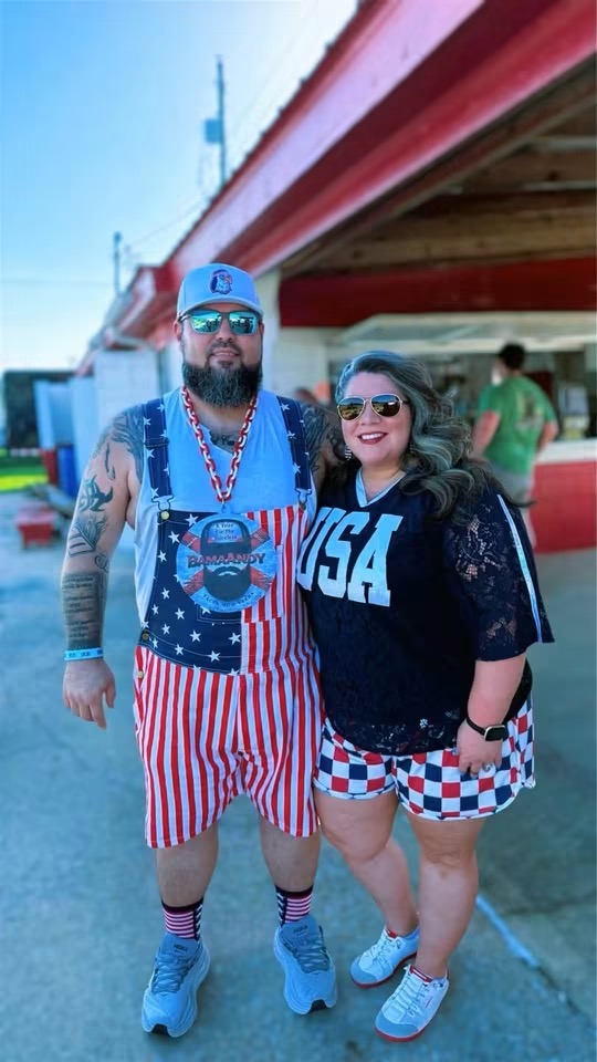 Two people standing outdoors, one wearing a blue cap, sunglasses, and patriotic shorts, the other in a black shirt with 'USA' and checkered shorts.