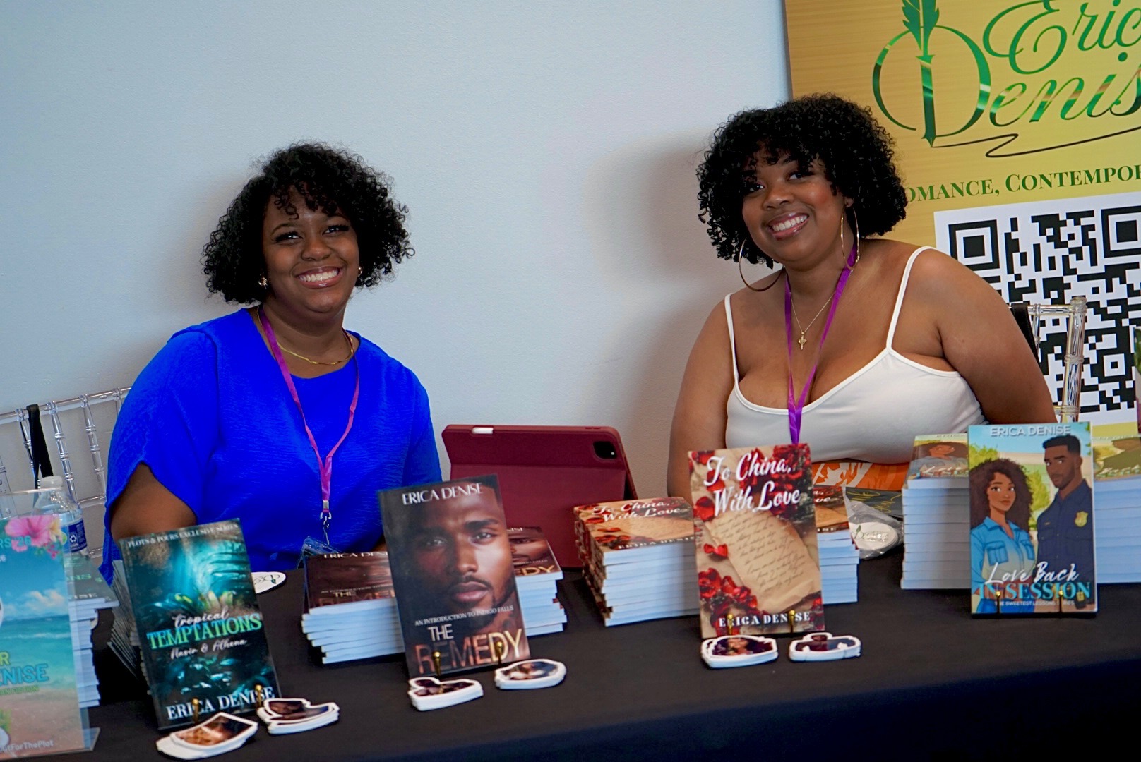 Two women sit behind a table with books and promotional materials, smiling at an event, with a yellow banner in the background.