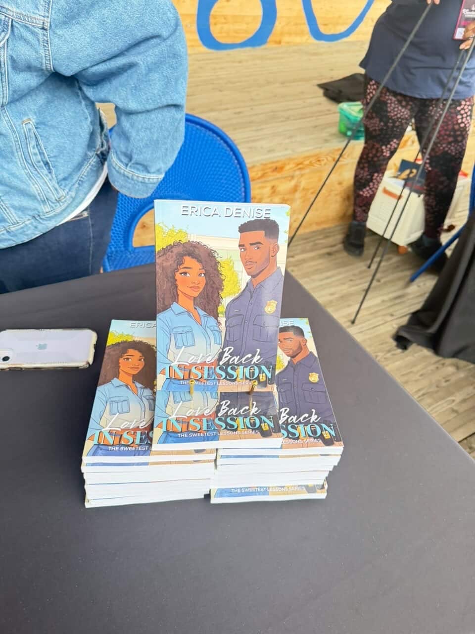 Stack of books with illustrated cover showing a girl and a boy, on a black table, with people and a wooden wall in background.