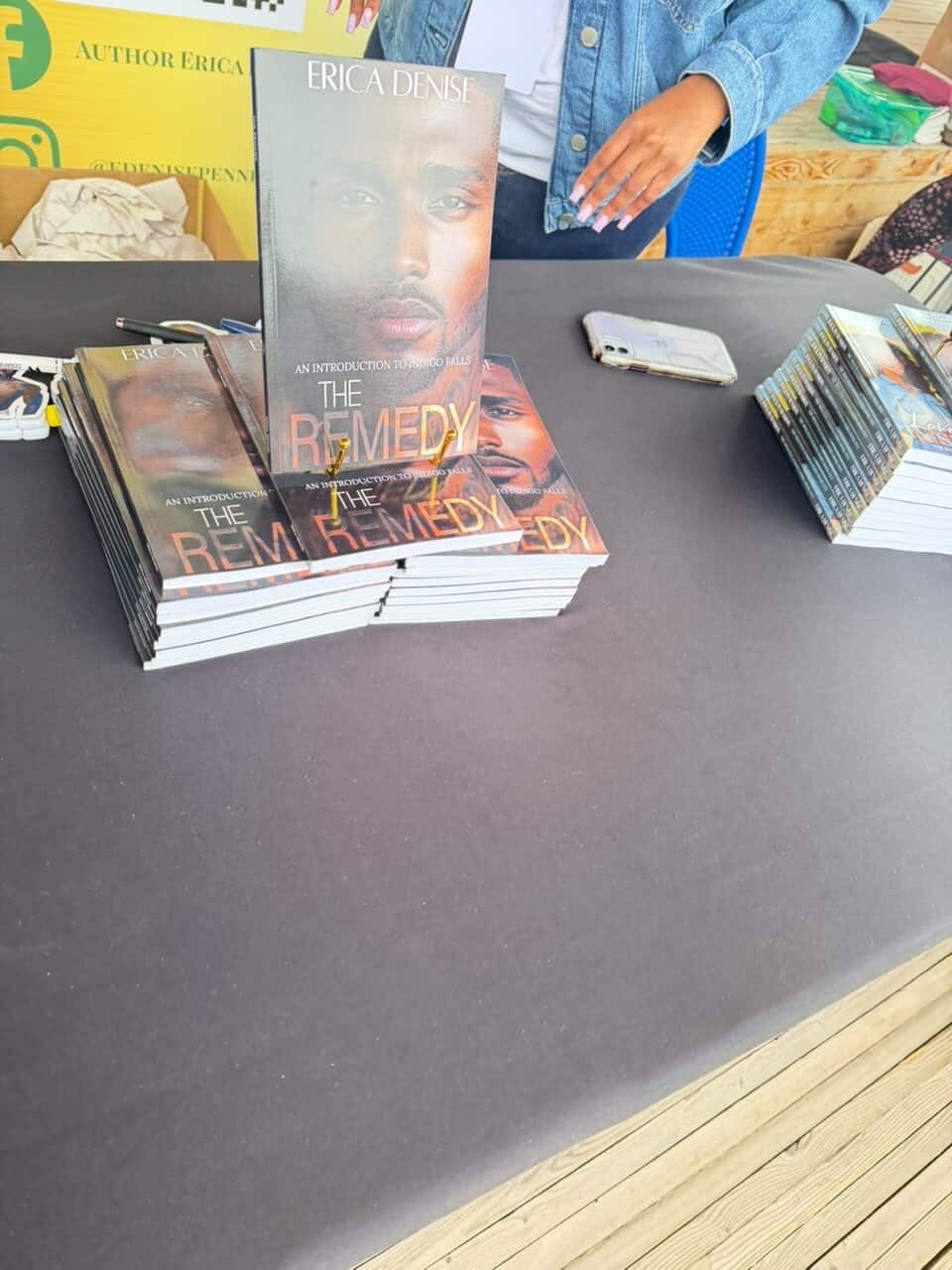 Stack of books titled 'The Remed' on a table with a person in a blue jacket behind, and other items nearby.