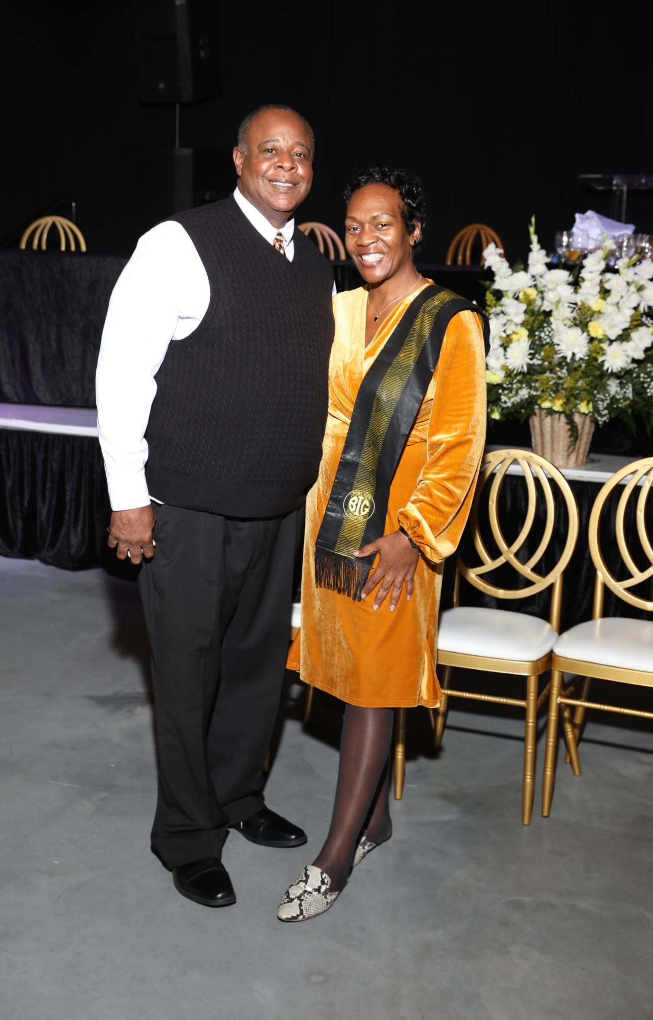 A man and woman stand together smiling at an event, with chairs and floral arrangements in the background.