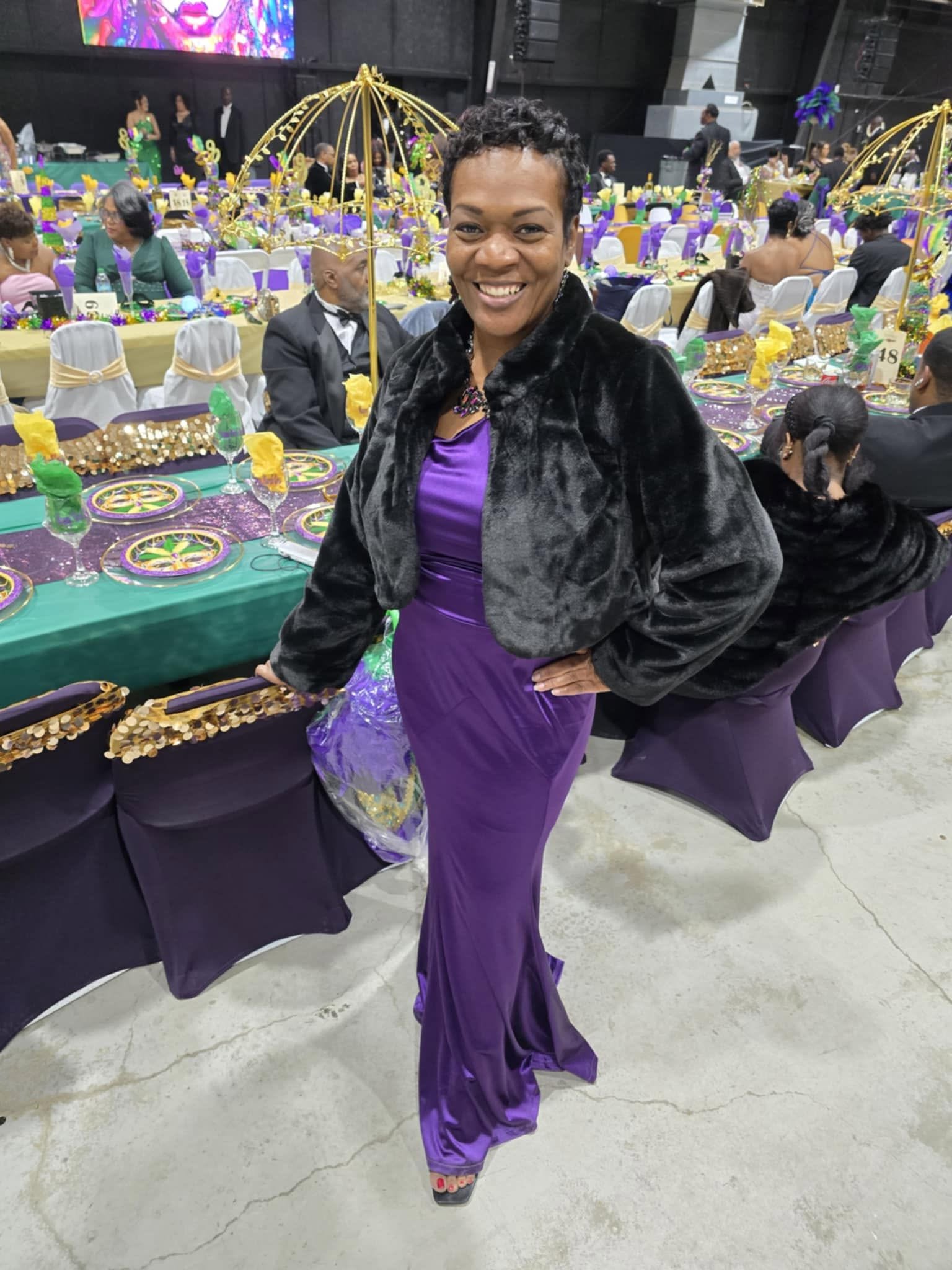 Woman in purple dress and black jacket smiling at event with decorated tables and guests.