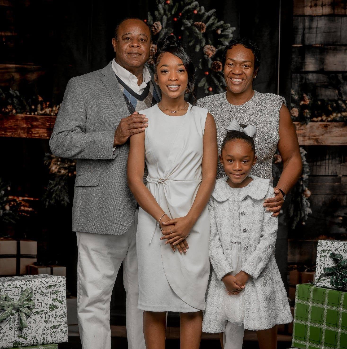 Family of four posing in front of a Christmas tree, smiling, with presents around them.