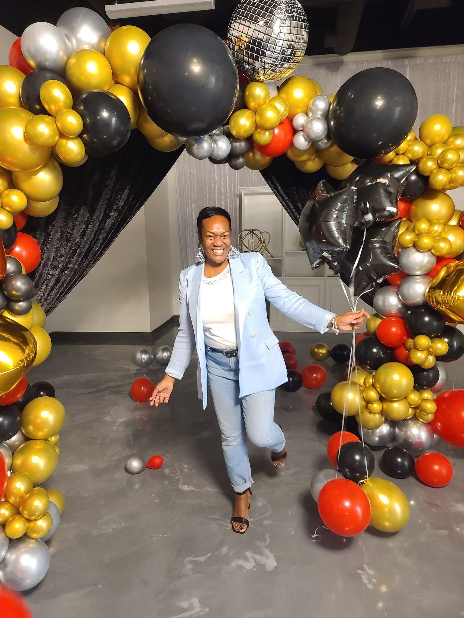 Person smiling and posing with arms outstretched in front of a balloon arch with black, gold, silver, and red balloons.