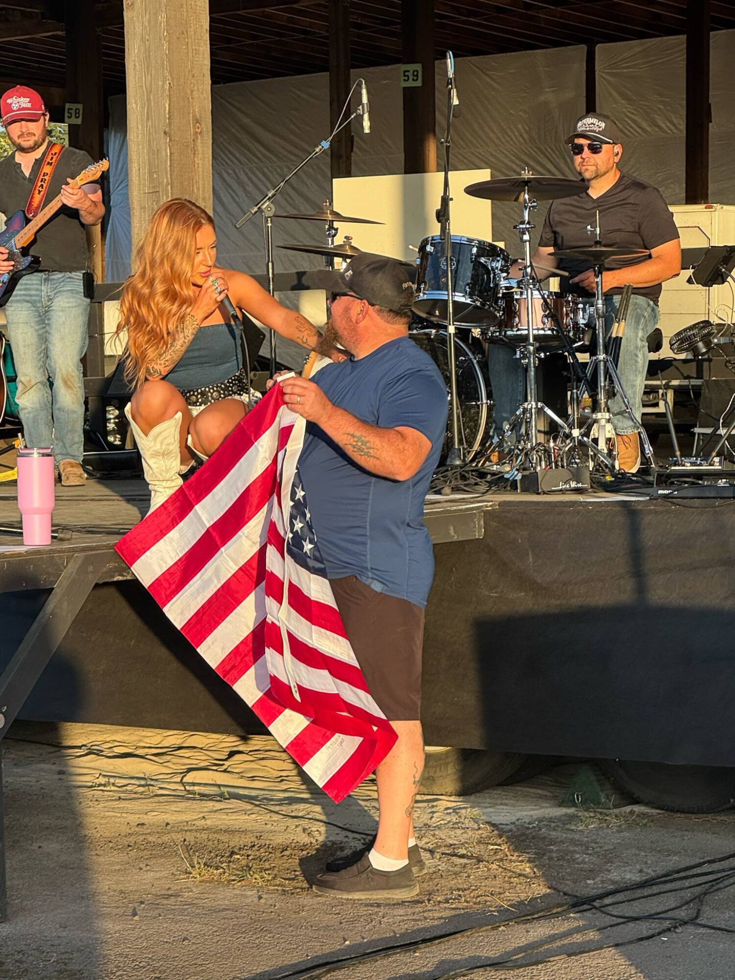 A woman and a man dance together outdoors, with a band playing on stage behind them. The woman has long hair and the man wears a hat. The man holds an American flag. Sunlight casts shadows on the ground.