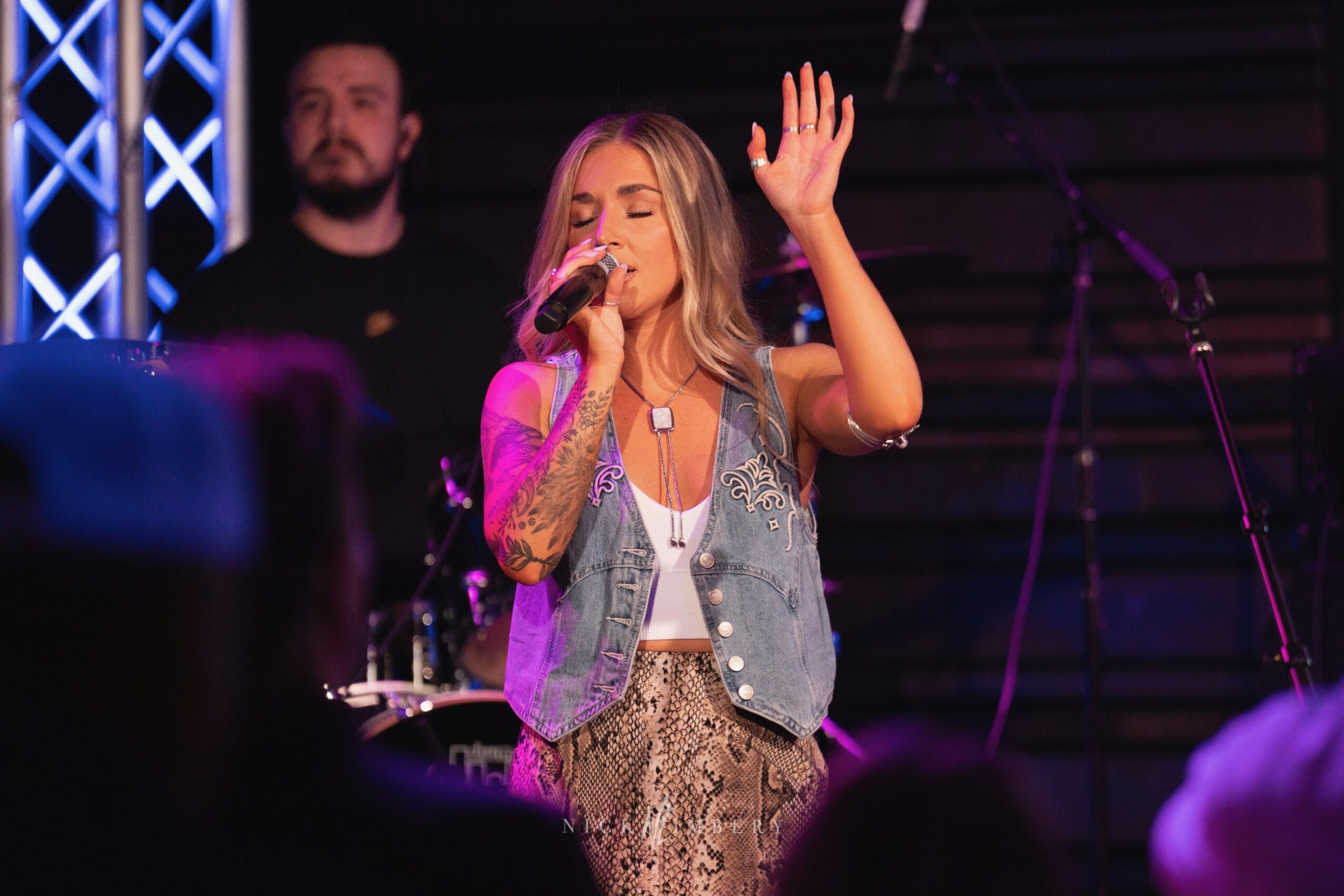A woman with long hair sings into a microphone with her eyes closed and hand raised on stage, with a man in the background.