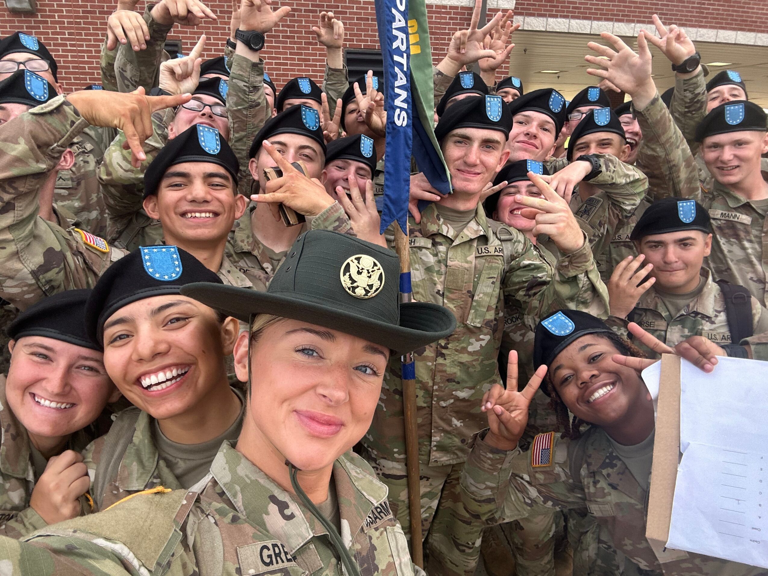 Group of smiling people in military uniforms, some making peace signs, gathered outdoors, with a woman wearing a black hat and glasses.