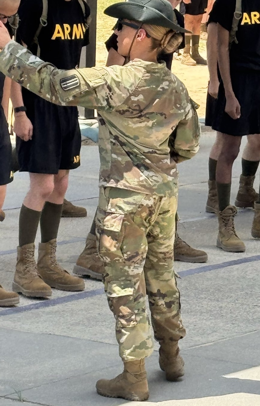 Child in camouflage military uniform with cap, standing outdoors among people in similar attire, raising arm.