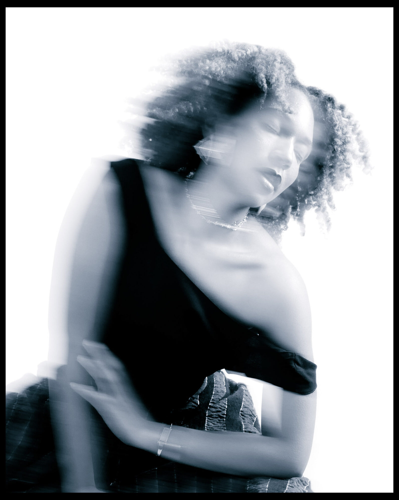 Blurry black-and-white portrait of a woman with curly hair, wearing a sleeveless top, with her eyes closed and hand near her face.