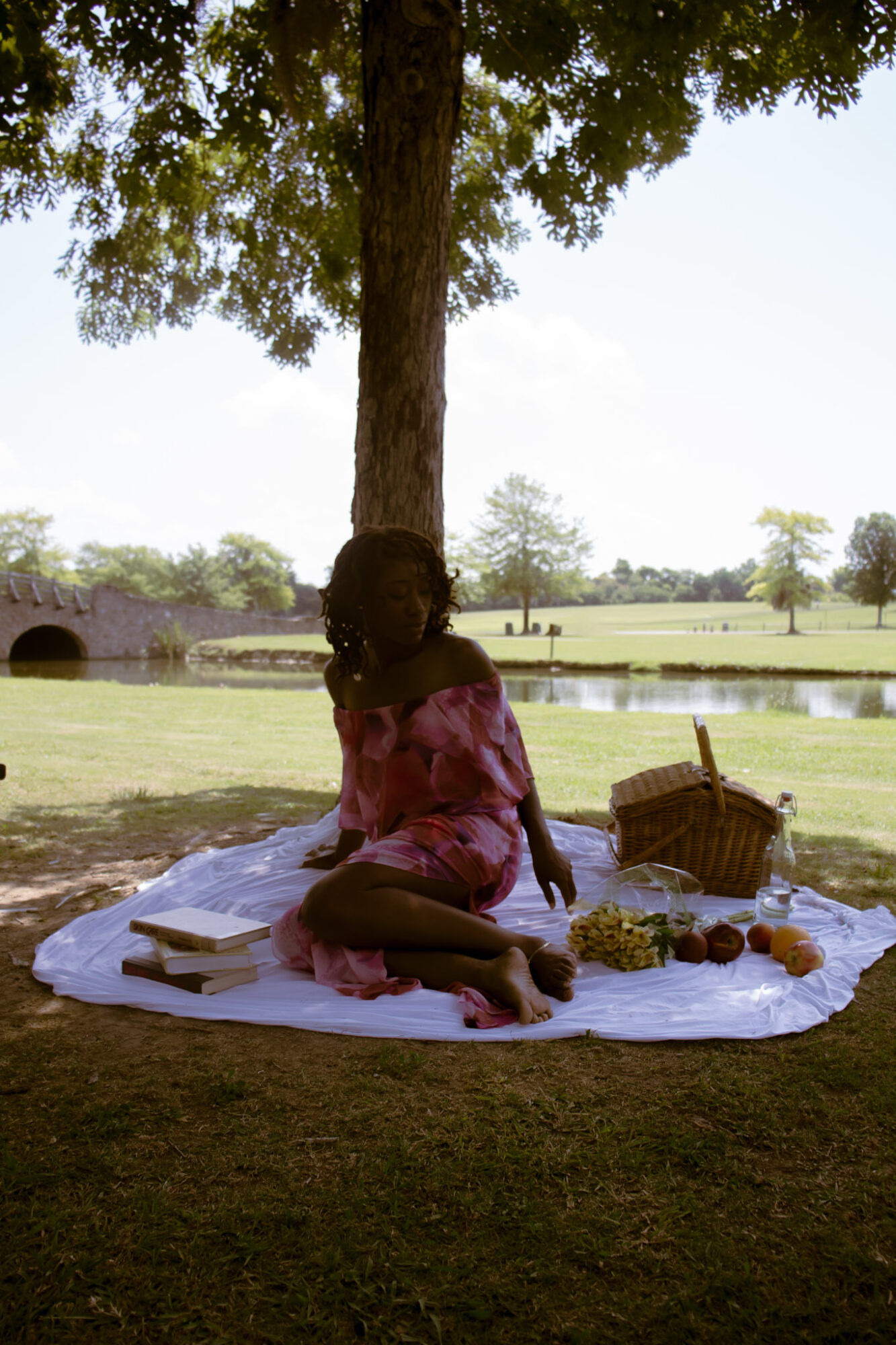 Child sitting on a white cloth under a tree with a basket and items nearby, near a body of water in a park.