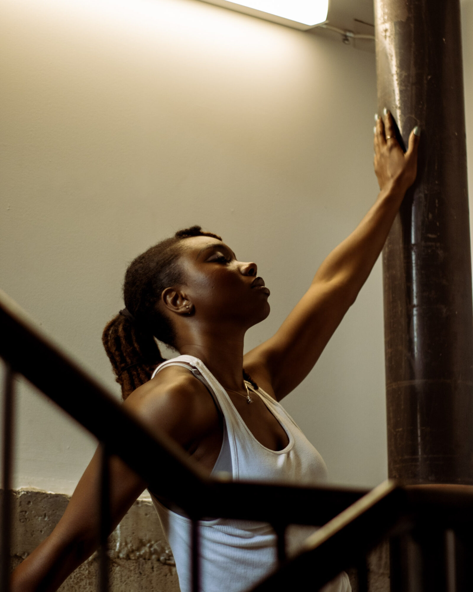 Woman with braided hair reaching upward, touching a pipe, in an indoor setting with a plain wall background.