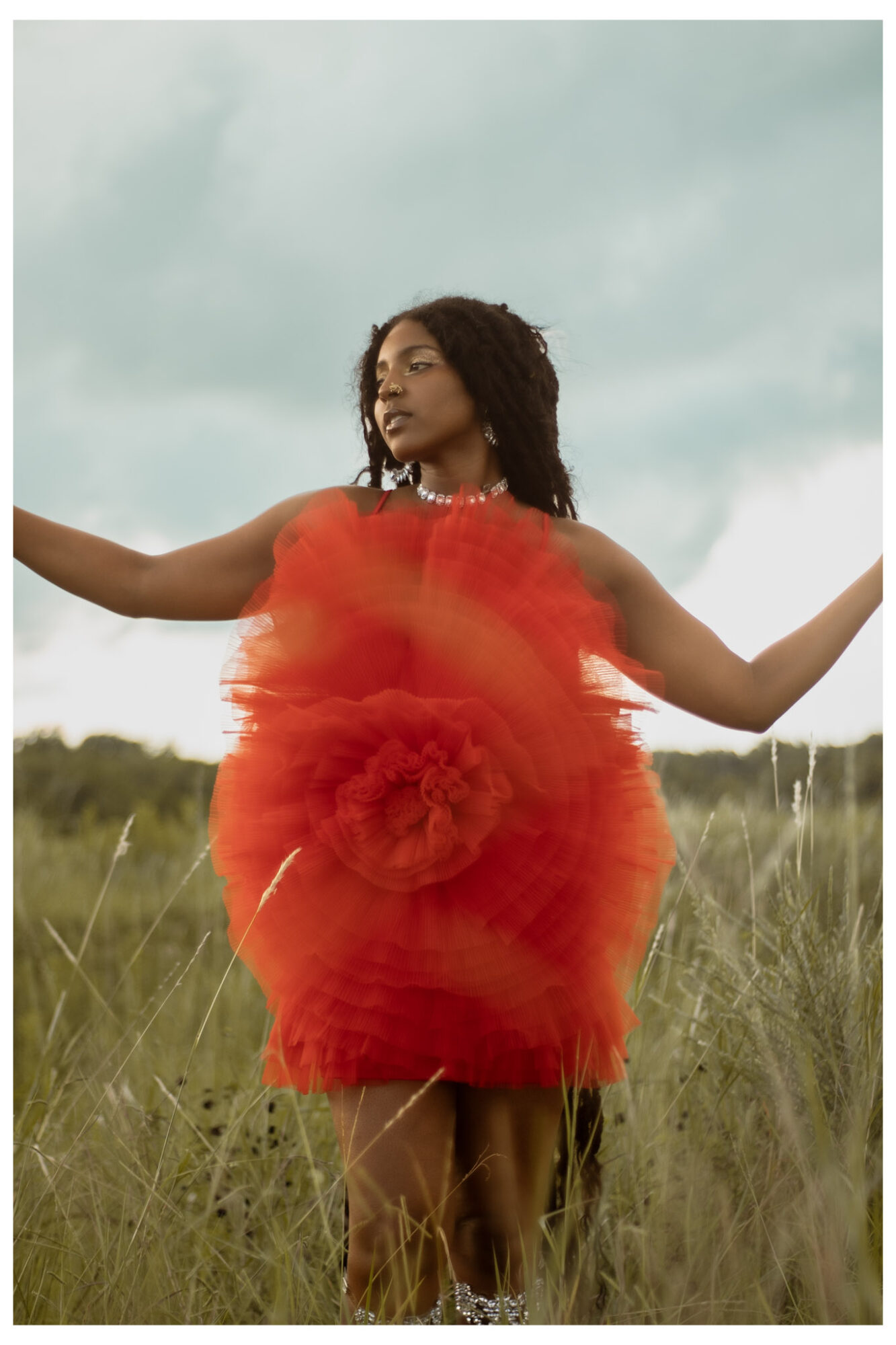 Woman standing in a grassy field wearing a bright red, fluffy dress with arms outstretched, sky in background.