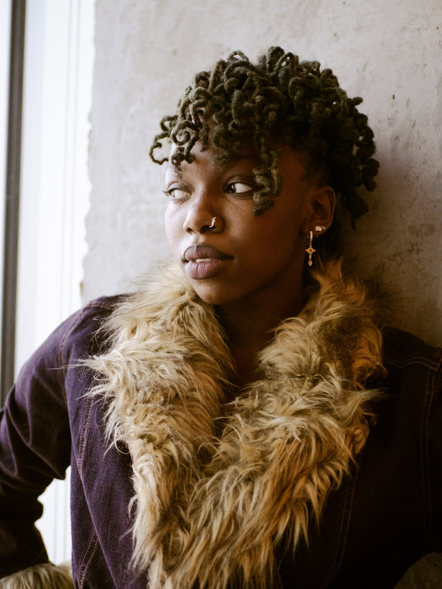 Woman with curly hair and fur collar looking to the side, standing against a textured wall.