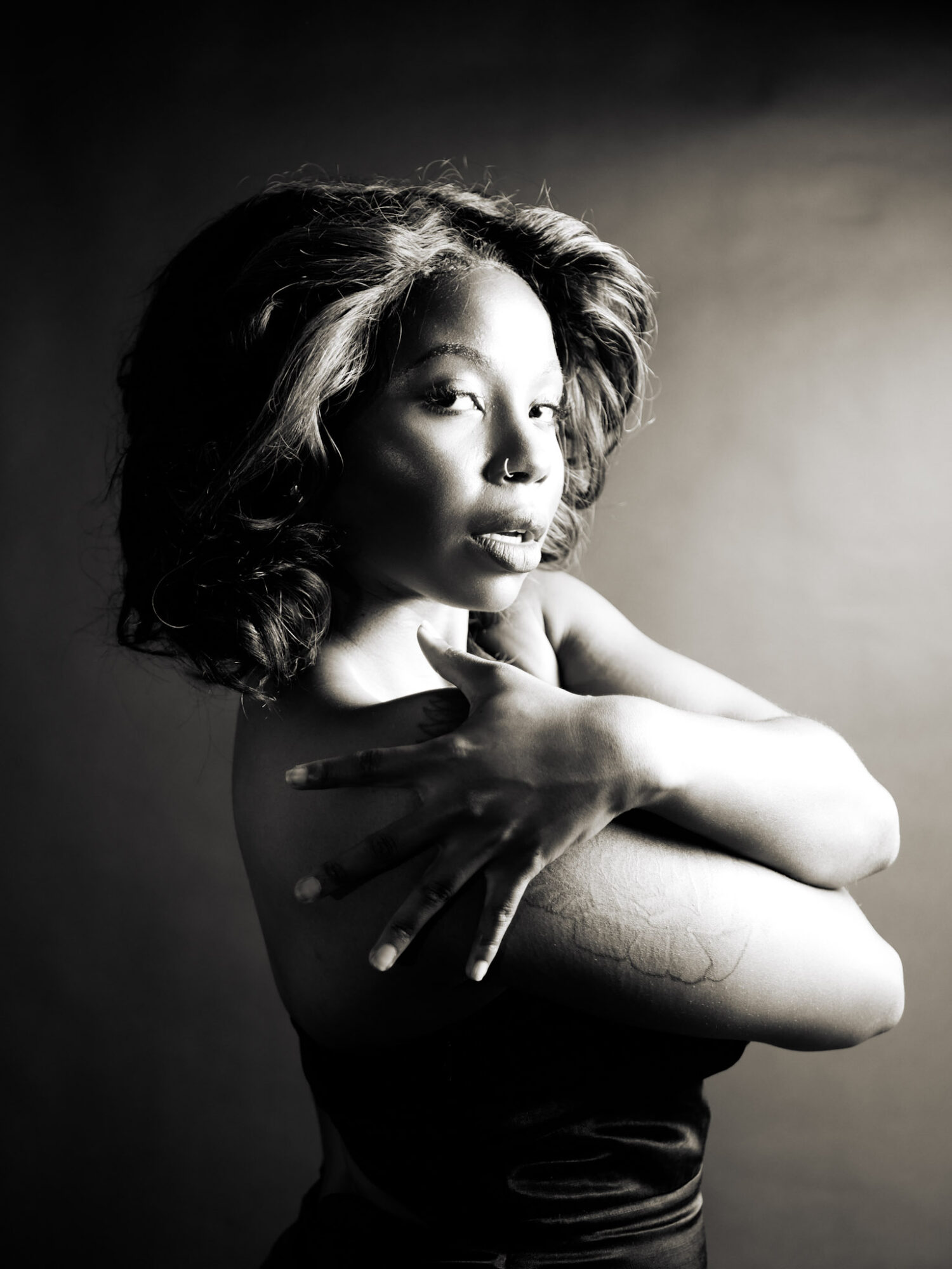 A woman with wavy hair poses with her arms crossed, looking to the side, in black and white.