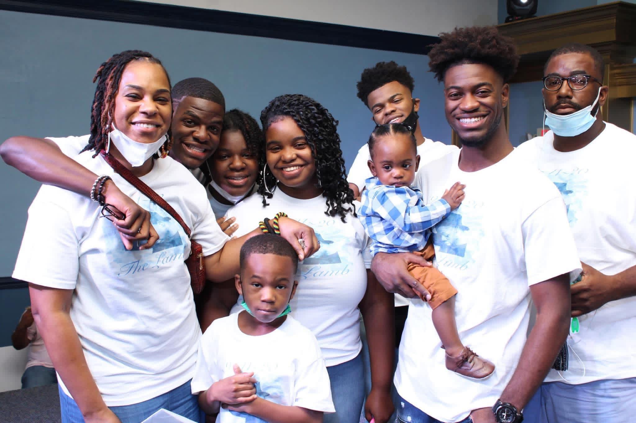 Group of smiling people, including children and adults, posing together indoors, some wearing masks around their necks.