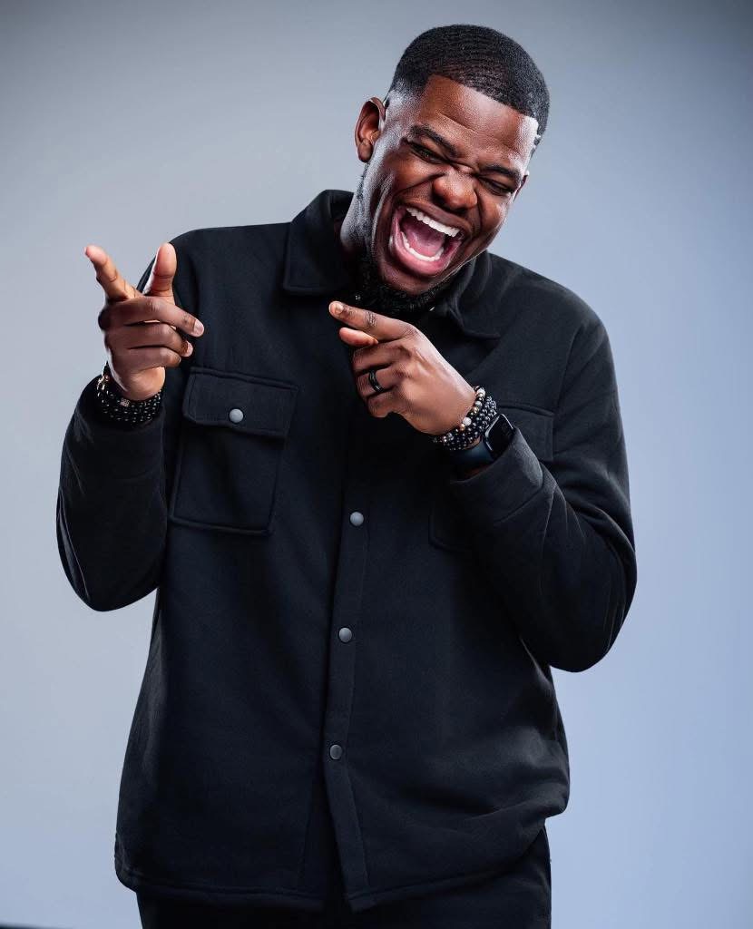 Person laughing with eyes closed, wearing a black jacket, pointing with both hands, against a light gray background.