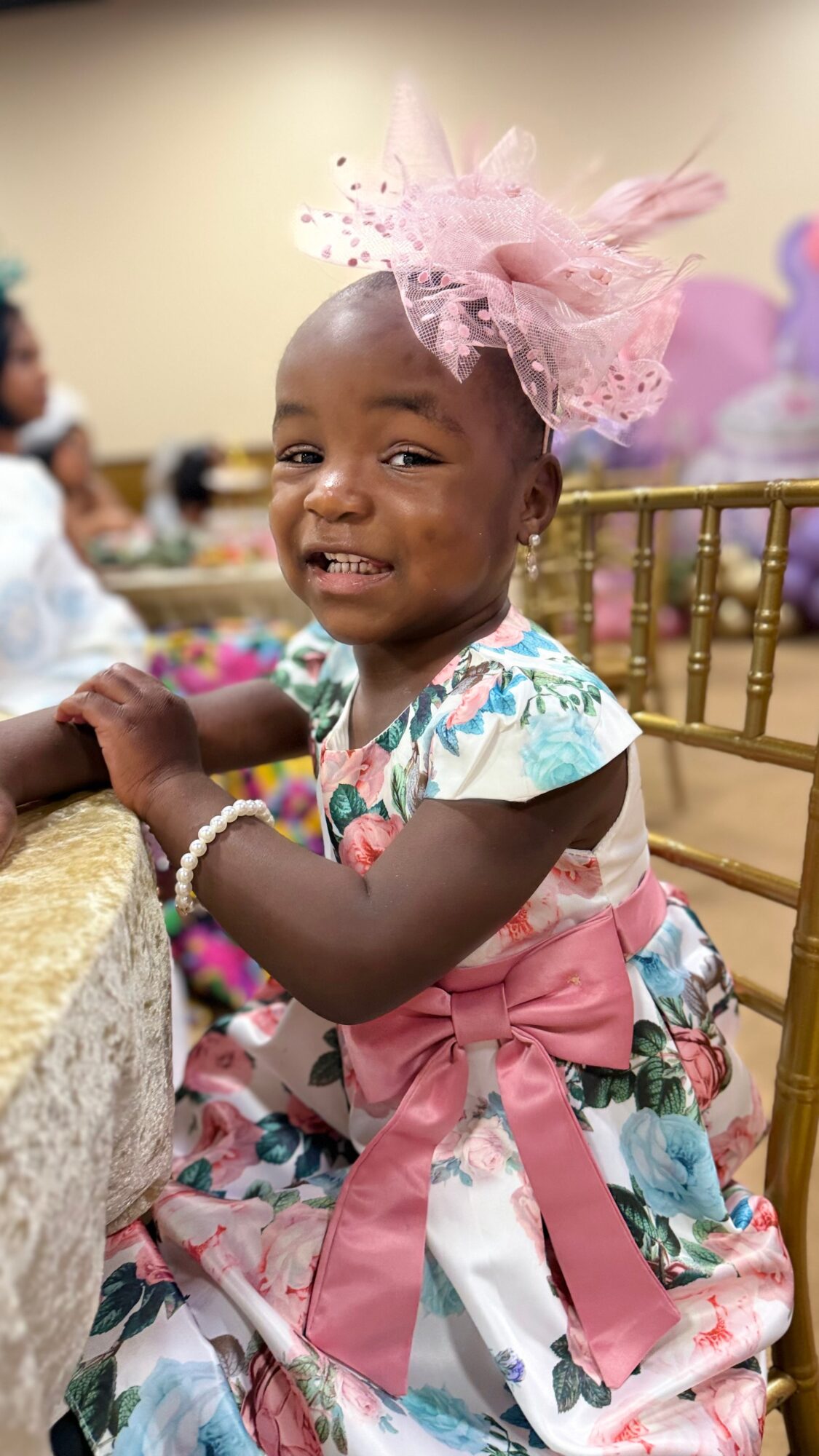 Young girl smiling, wearing a floral dress with pink ribbon, pink tulle headpiece, sitting at a table with gold chairs.