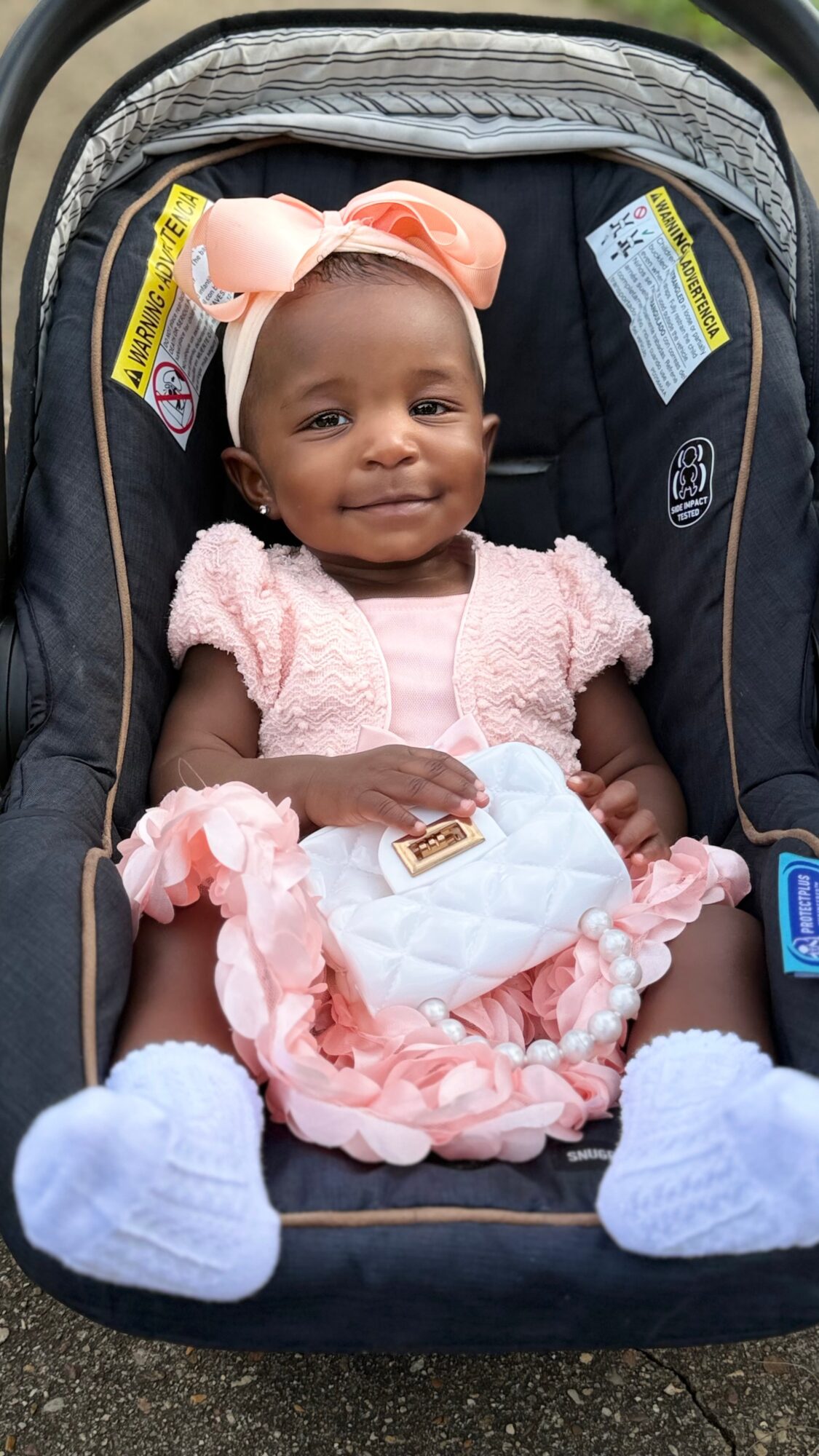 Smiling baby girl in a car seat wearing a pink dress, bow, and white socks, holding a small white quilt.