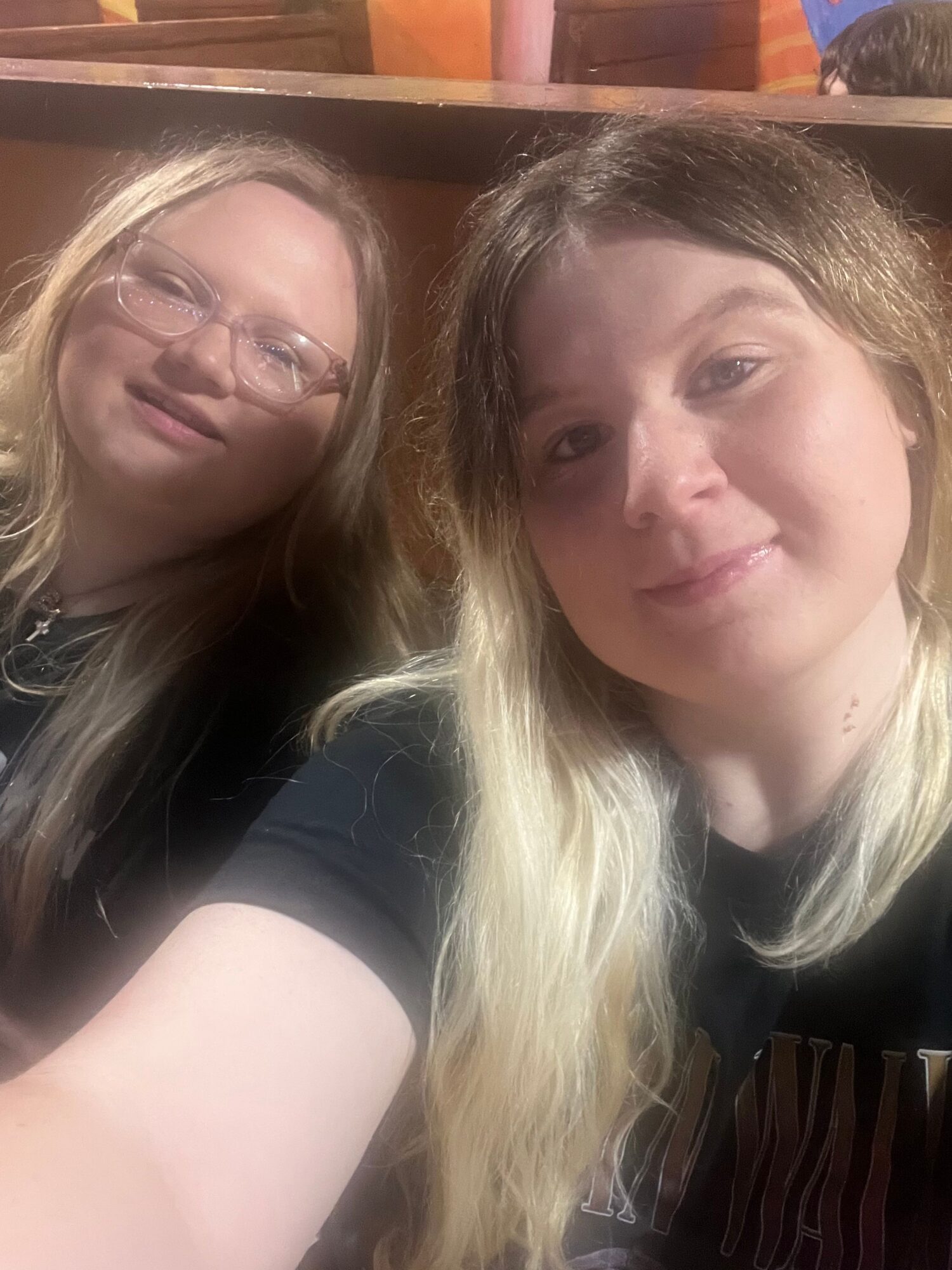 Two women with long hair, one with glasses, smiling and looking at the camera, in an indoor setting.