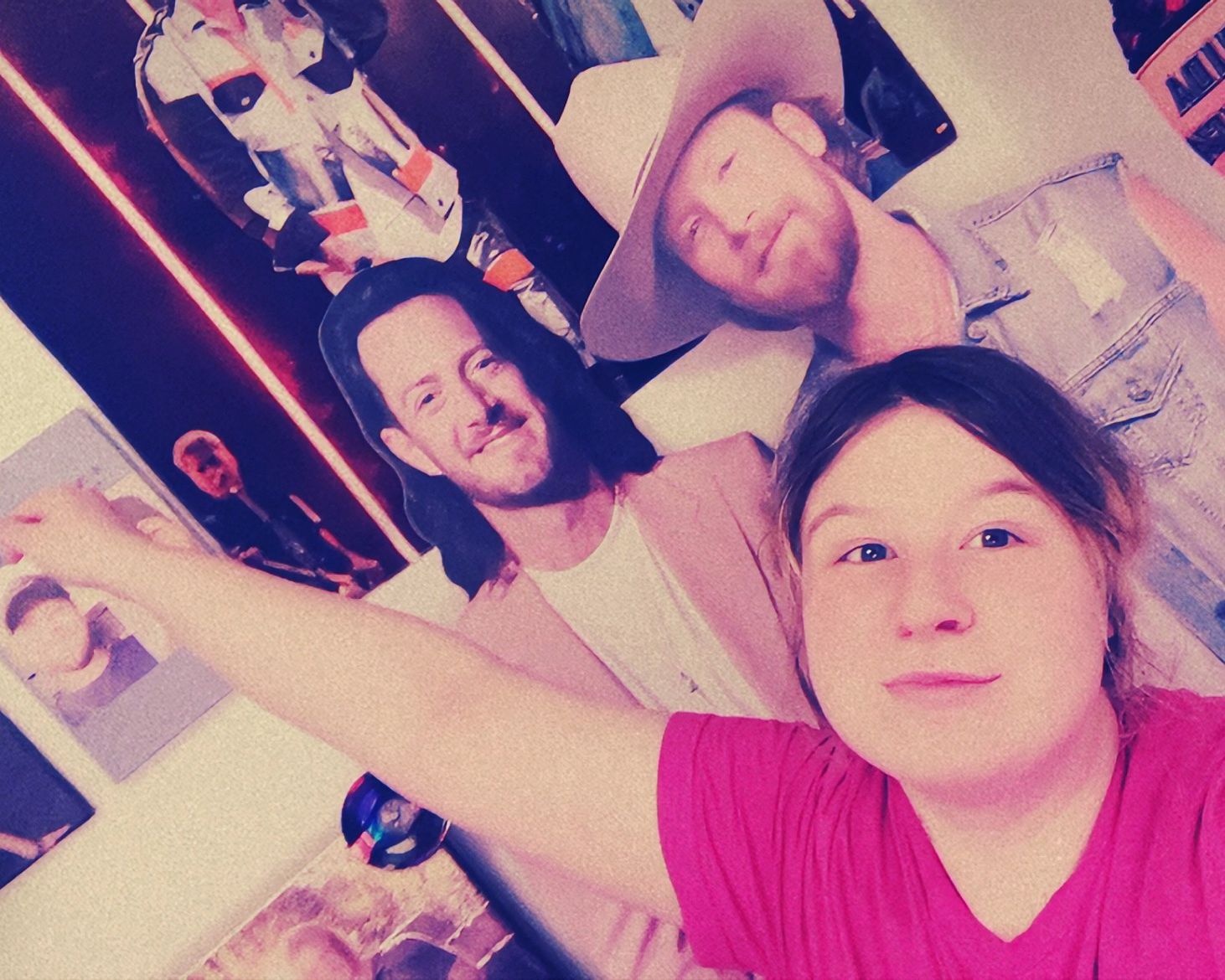 Three people smiling, one wearing a pink shirt, another with a hat, in a room with artwork on the wall.
