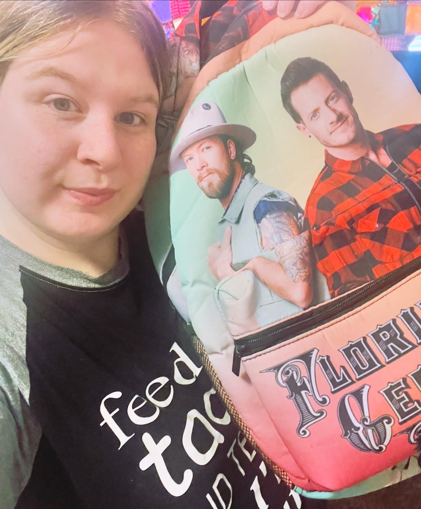 Close-up of a woman with light brown hair, wearing a black shirt with white text, standing next to a colorful backpack featuring two men, one in a hat and the other in a red plaid shirt.