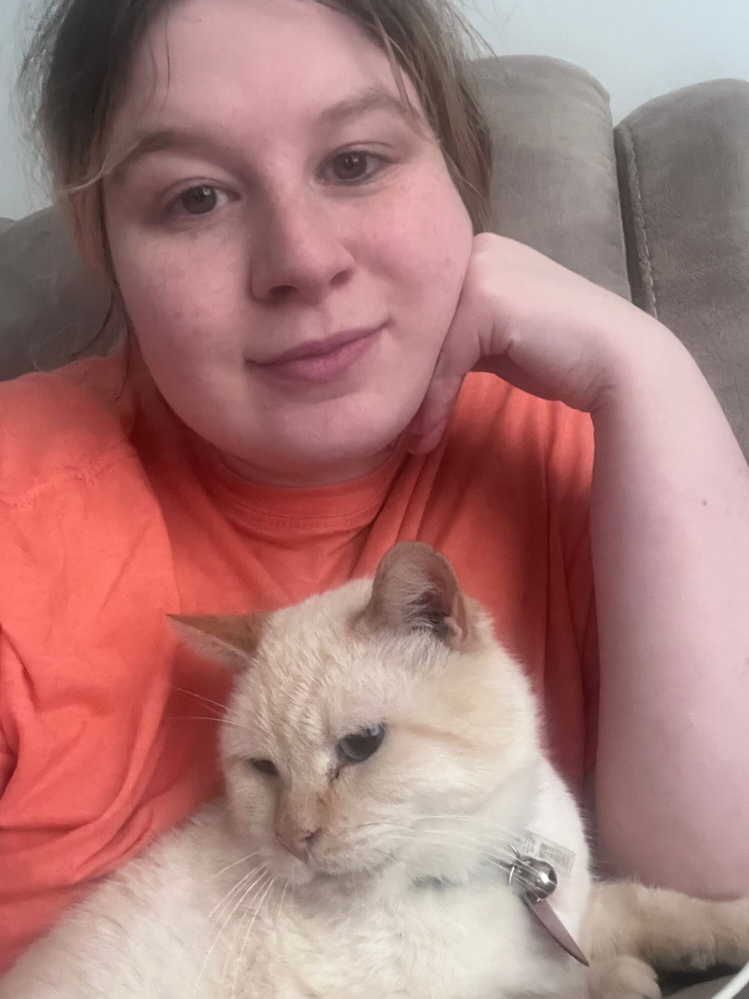 Woman lying on a couch with a cream-colored cat, both looking at the camera, woman wearing an orange shirt.