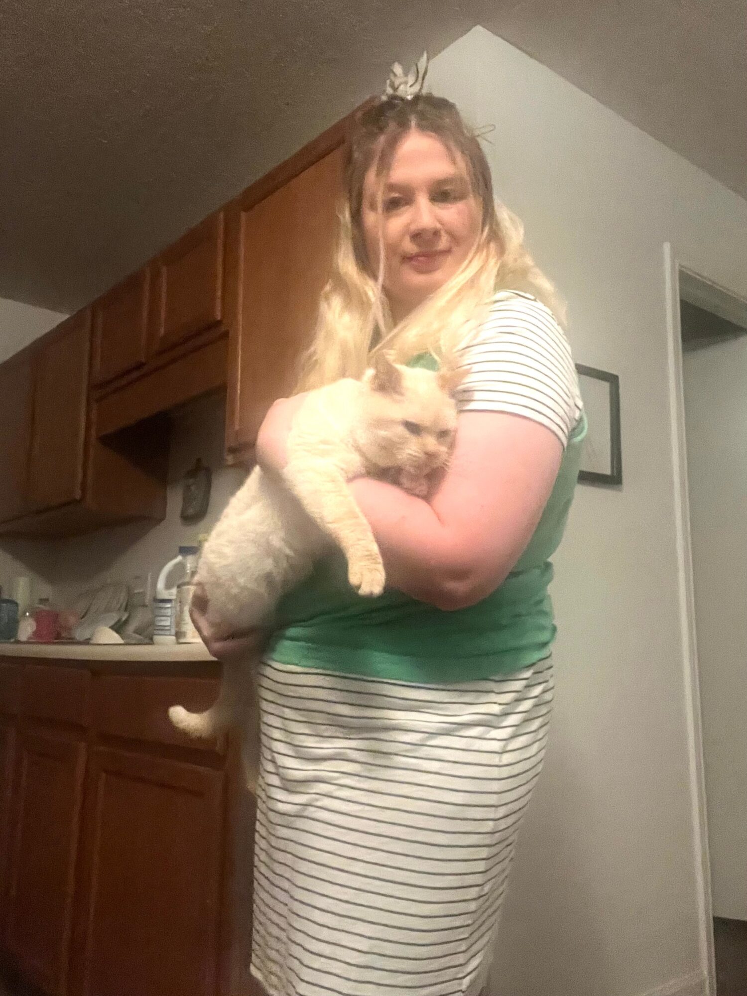 Person with long blonde hair holding a light-colored cat in a kitchen, smiling at the camera.