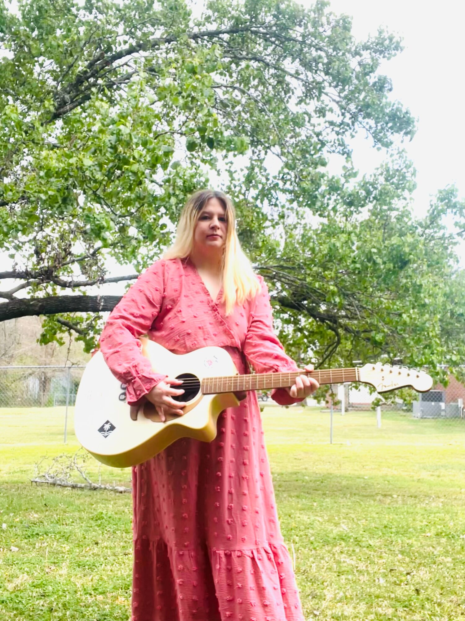 Woman in pink dress holding a white acoustic guitar outdoors with trees and grass in background.