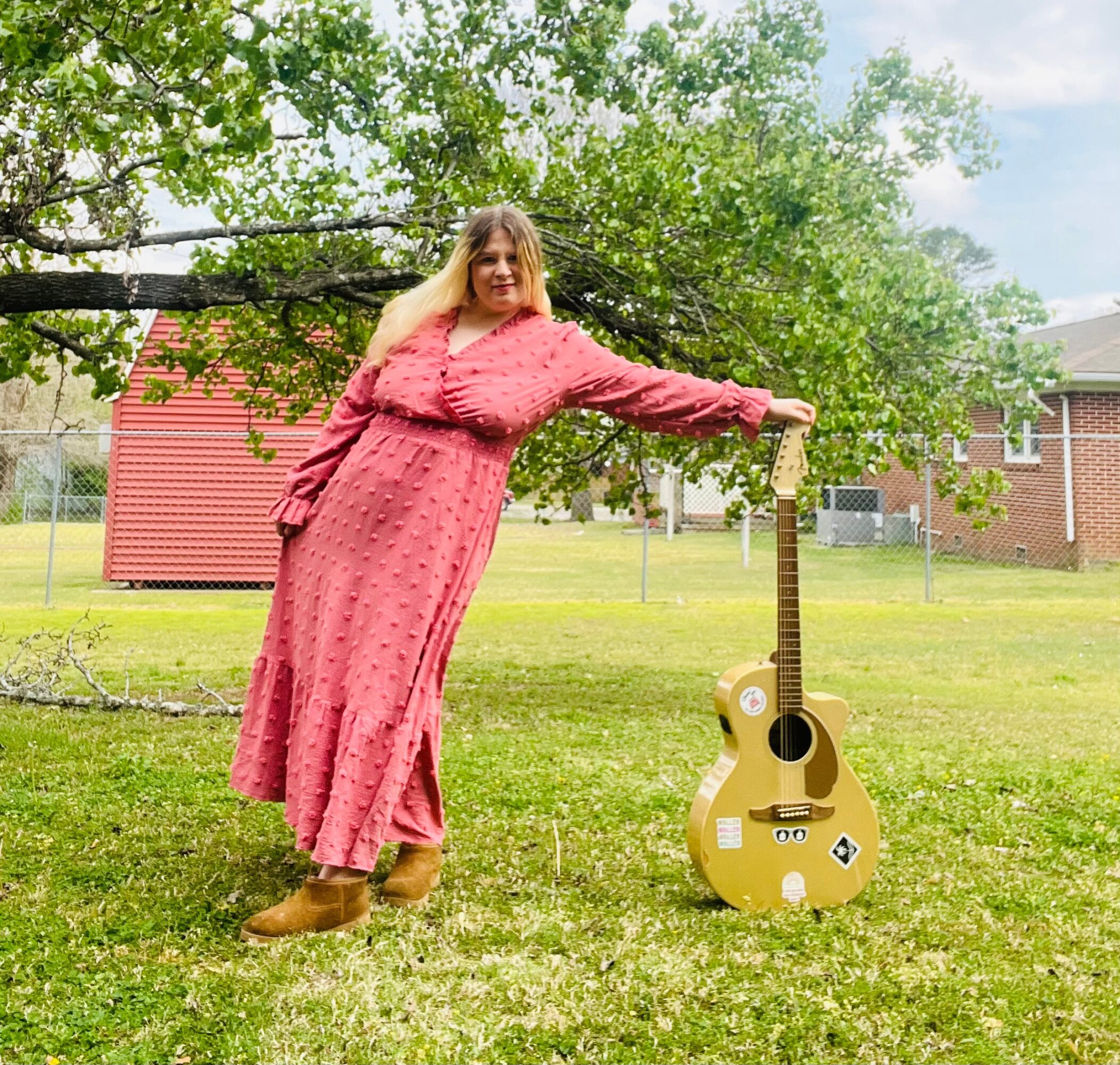 Woman in pink dress holding a tree branch, standing next to a decorated guitar outdoors, with grass and trees in background.