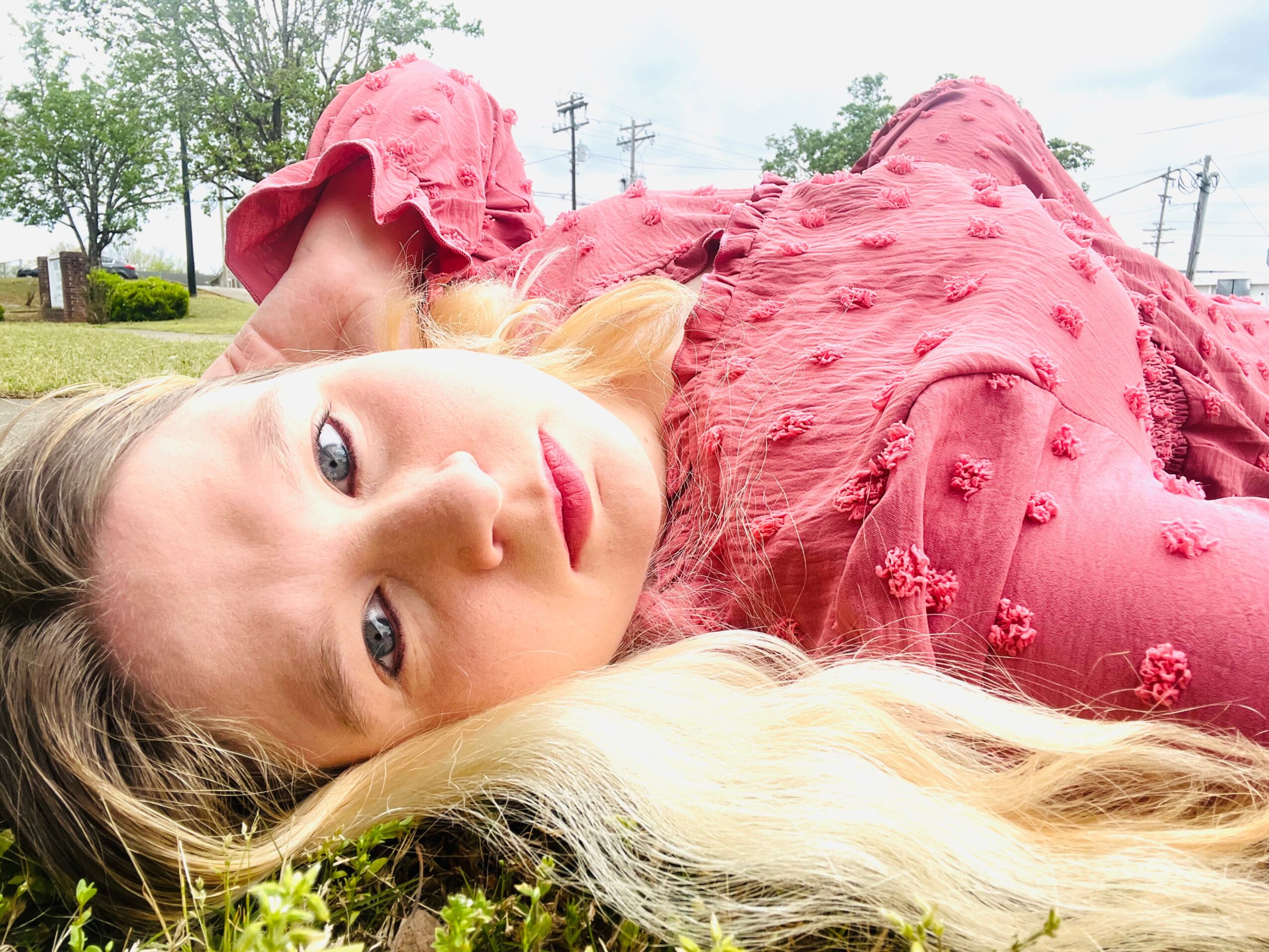 Young woman lying outdoors on grass, wearing a pink dress, with blonde hair and blue eyes, looking at the camera.