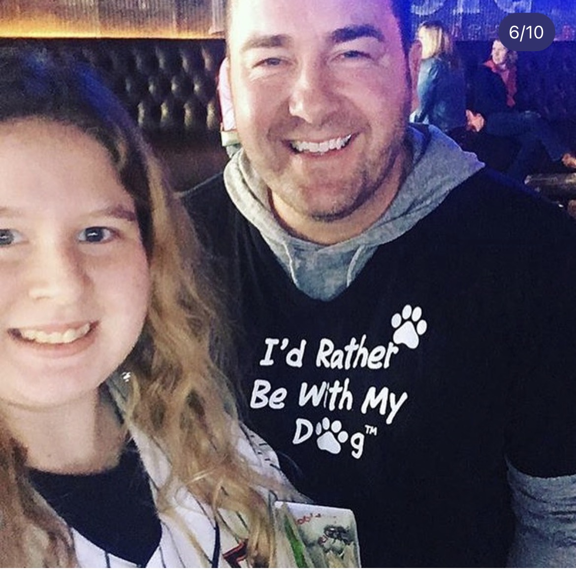 Two people smiling, one wearing a black shirt with text and a paw print, in a social setting.