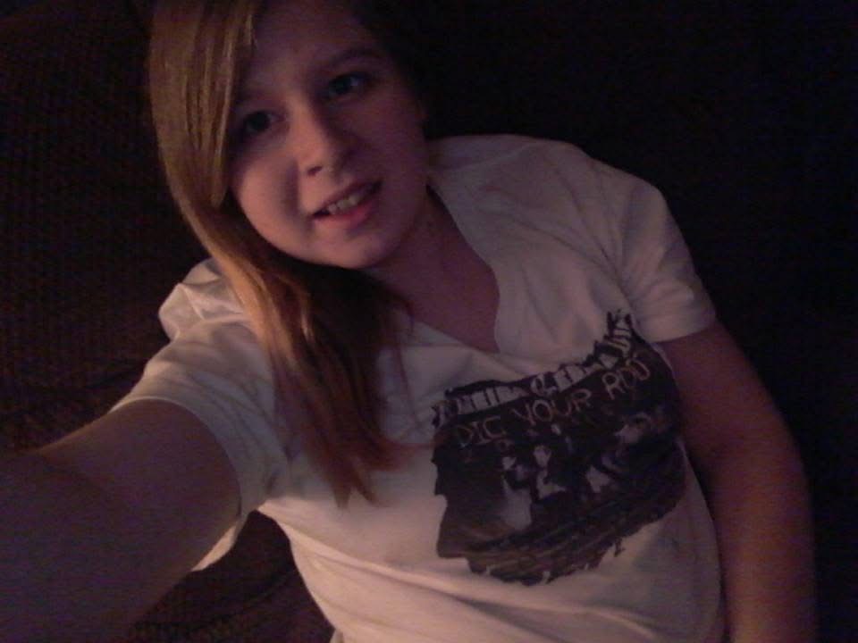 Young girl with long hair smiling, wearing a white T-shirt, in a dark setting.