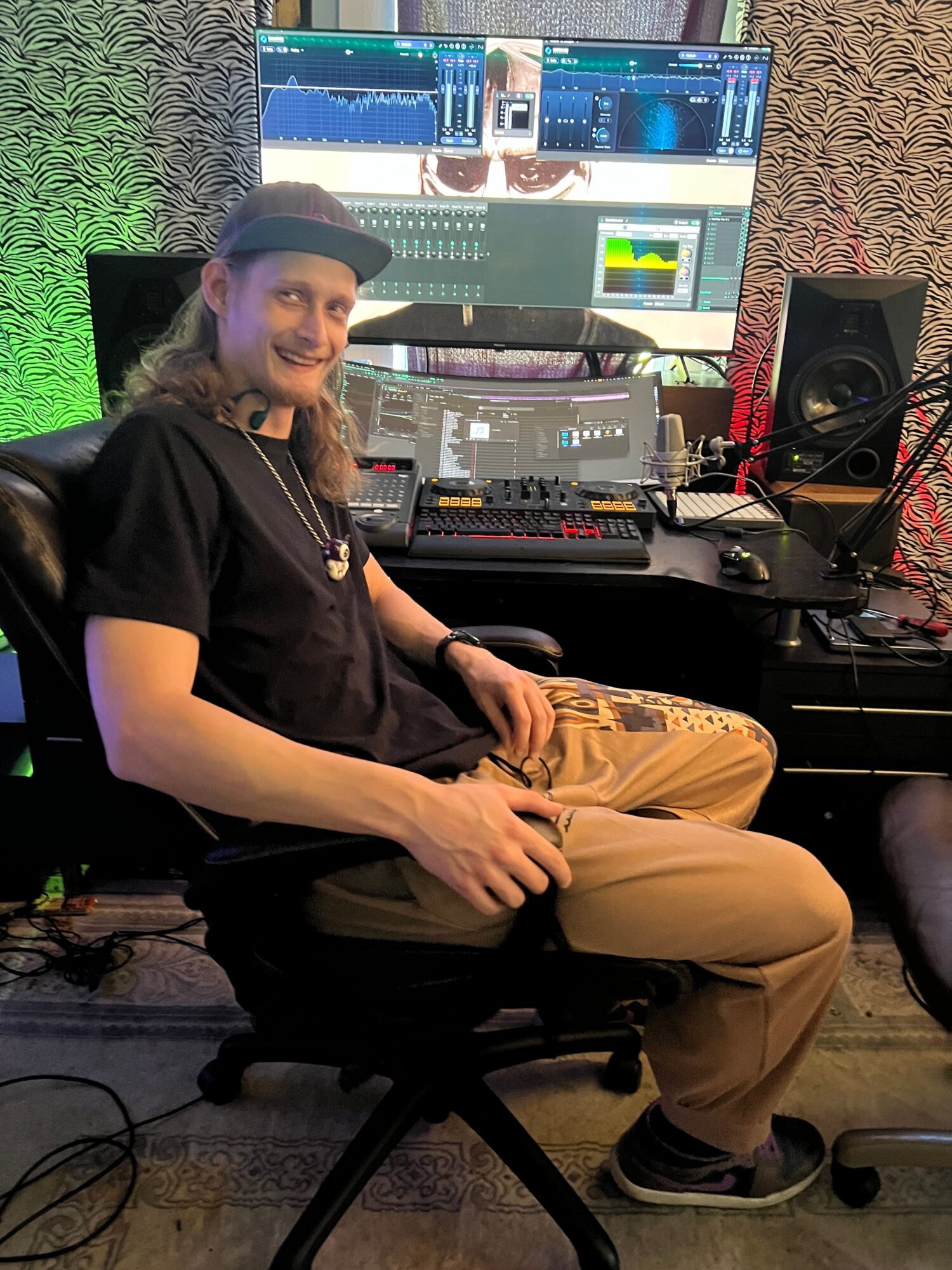 Person sitting in front of DJ equipment and computer screens, wearing a cap and casual clothing, in a music studio.