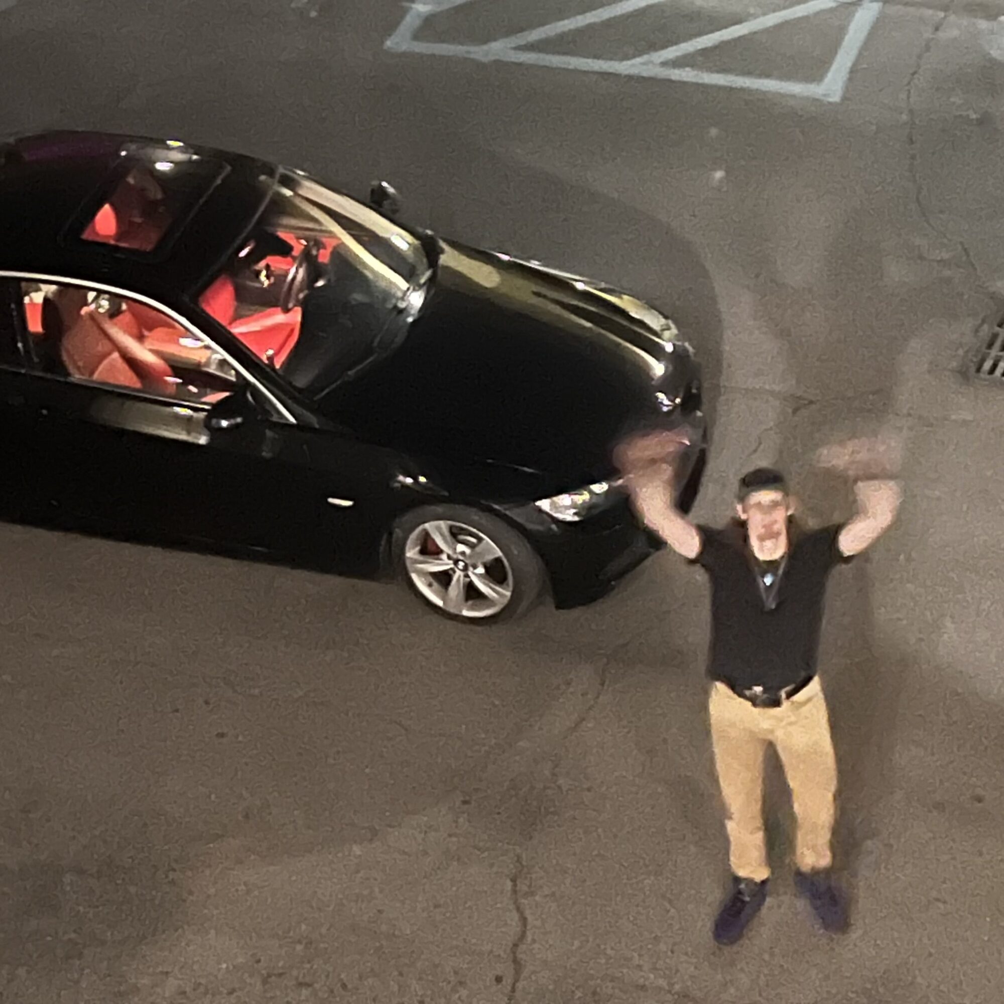 Person standing in parking lot with arms raised, black car parked nearby, night setting, visible parking lines.