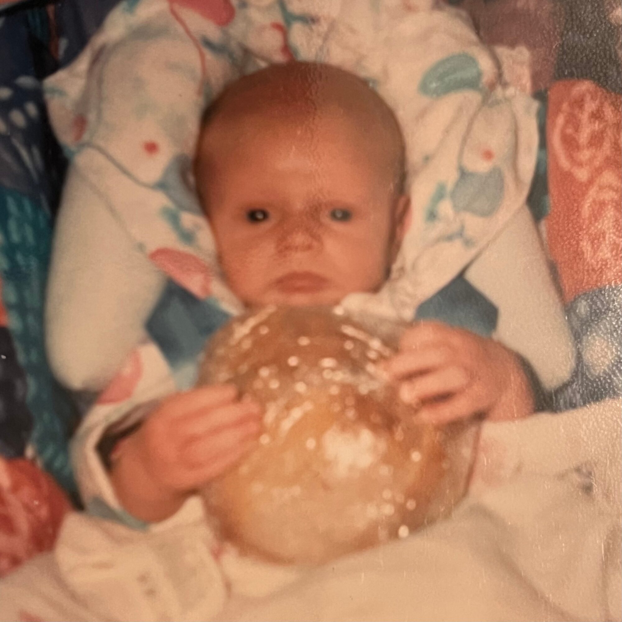 Baby lying in a bed holding a large bread roll, with a blanket and patterned bedding visible.