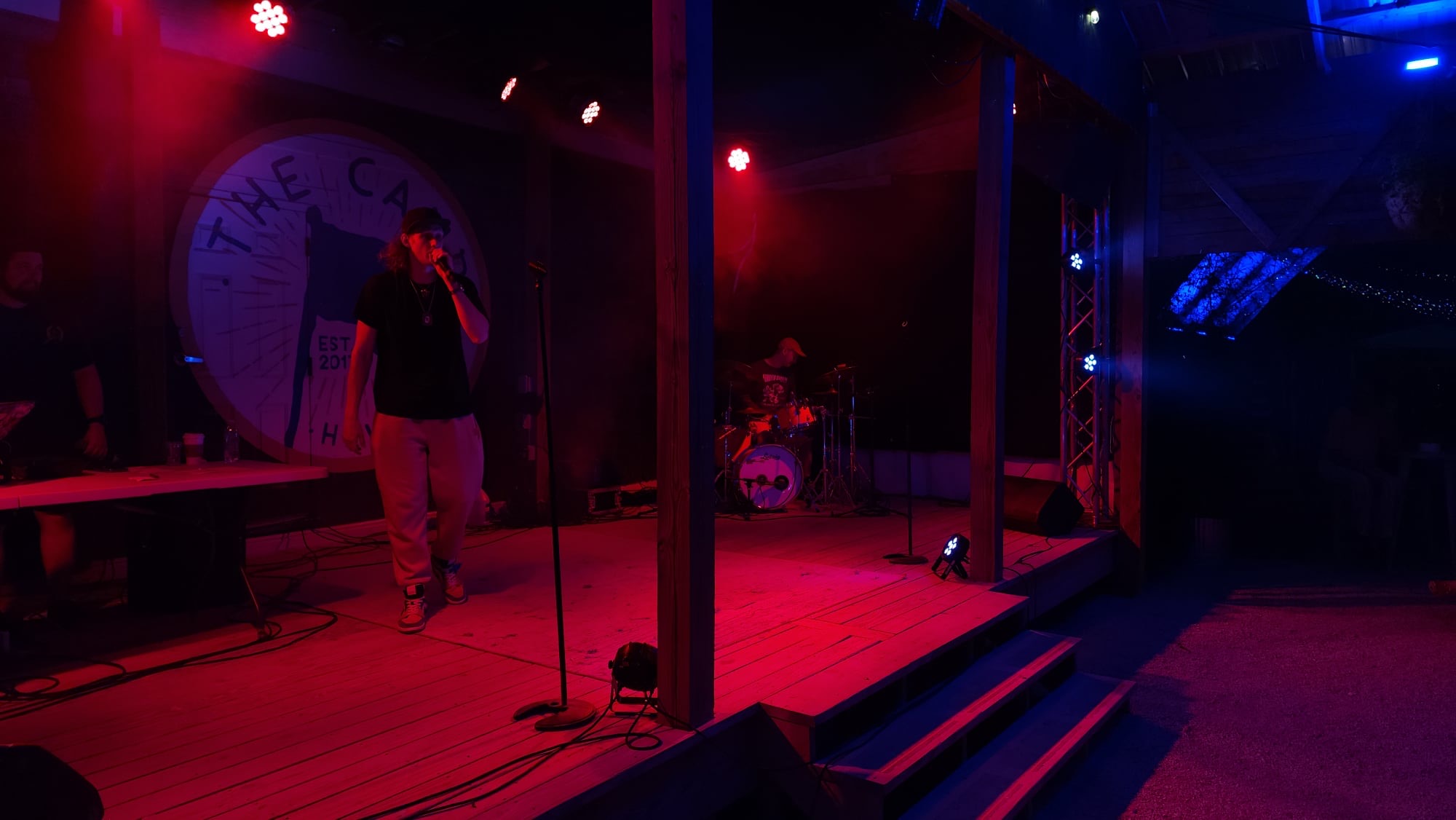Stage with microphone stand, person preparing, drum set in background, dim lighting, red and blue lights, wooden floor, steps to stage
