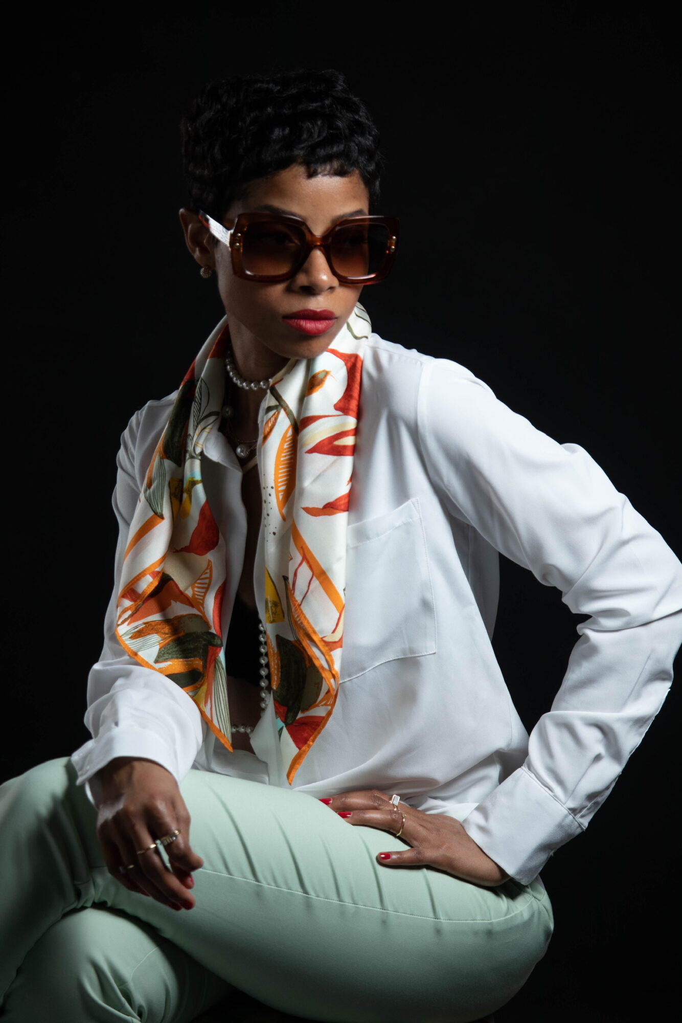 Woman with short curly hair wearing sunglasses, a white shirt, and a colorful scarf, posing against a black background.