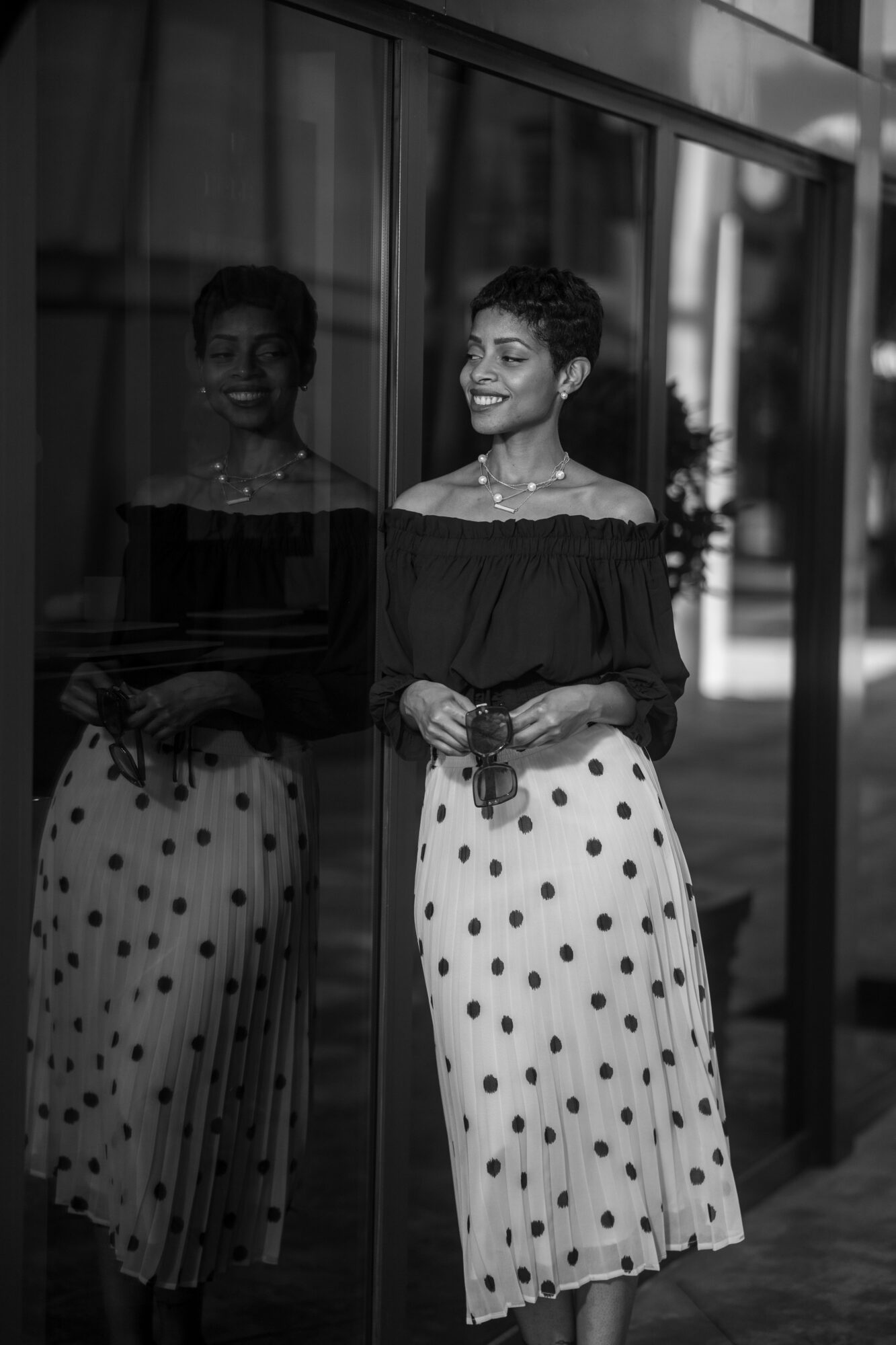 Woman with short hair smiling, standing next to a glass wall, her reflection visible, wearing a polka dot skirt and off-shoulder top.