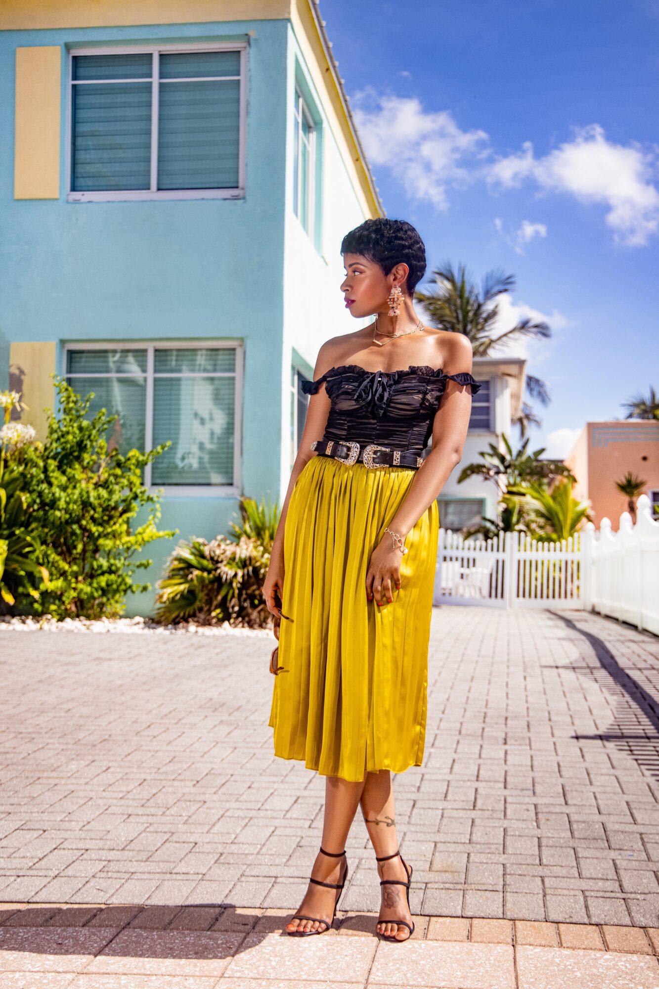 Woman in black top and yellow skirt standing outdoors on paved path with building and palm trees in background.