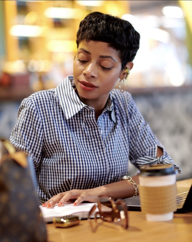 Woman with short black hair, wearing a checkered shirt, sitting at a table with a coffee cup and notebook.
