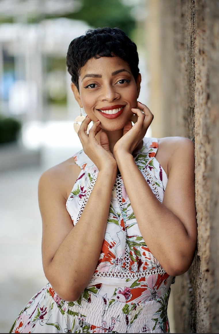 Smiling woman with short dark hair, wearing a colorful sleeveless dress, standing outdoors near a textured wall.