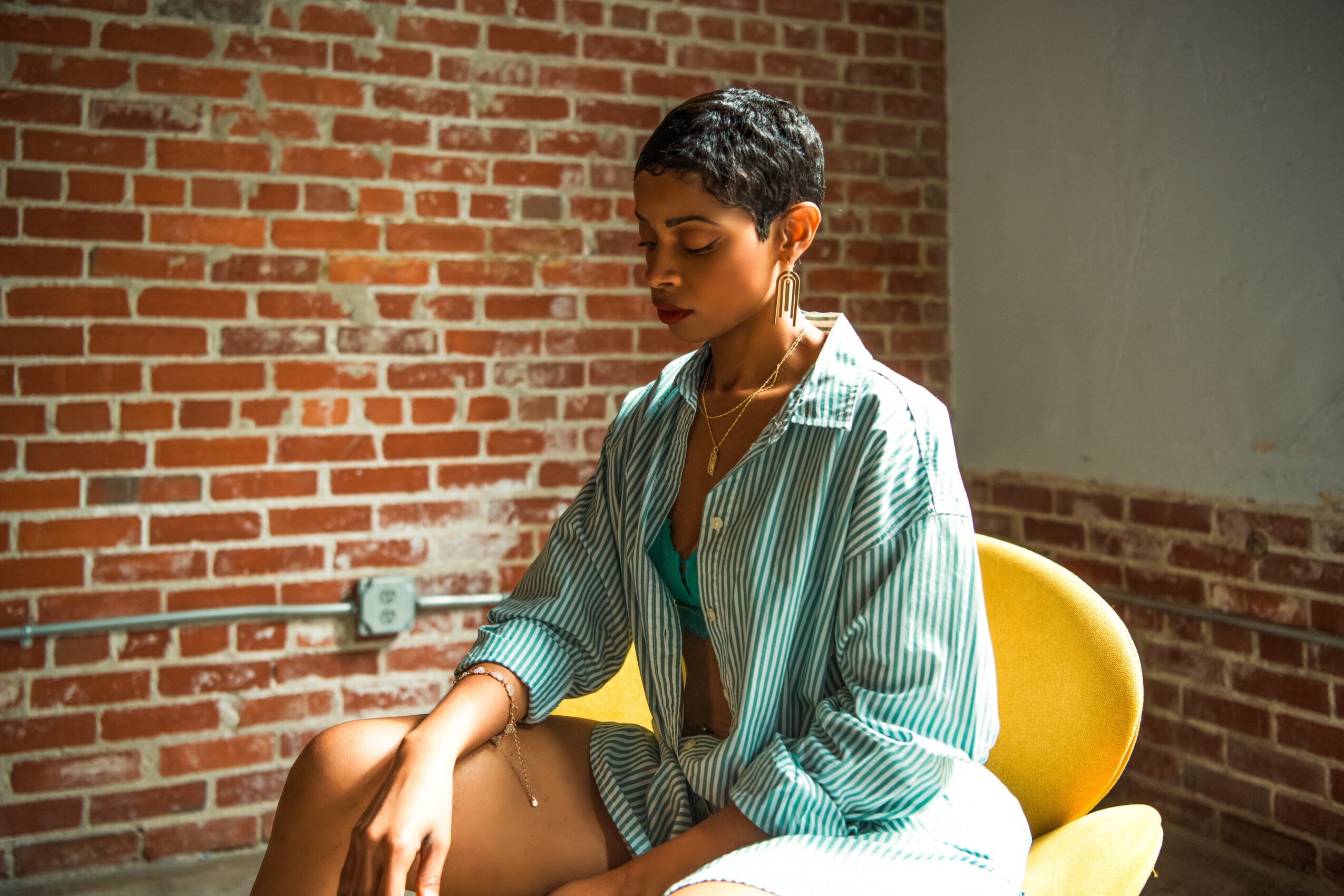 Woman with short hair sitting on a yellow chair against a brick wall, looking down with a contemplative expression.