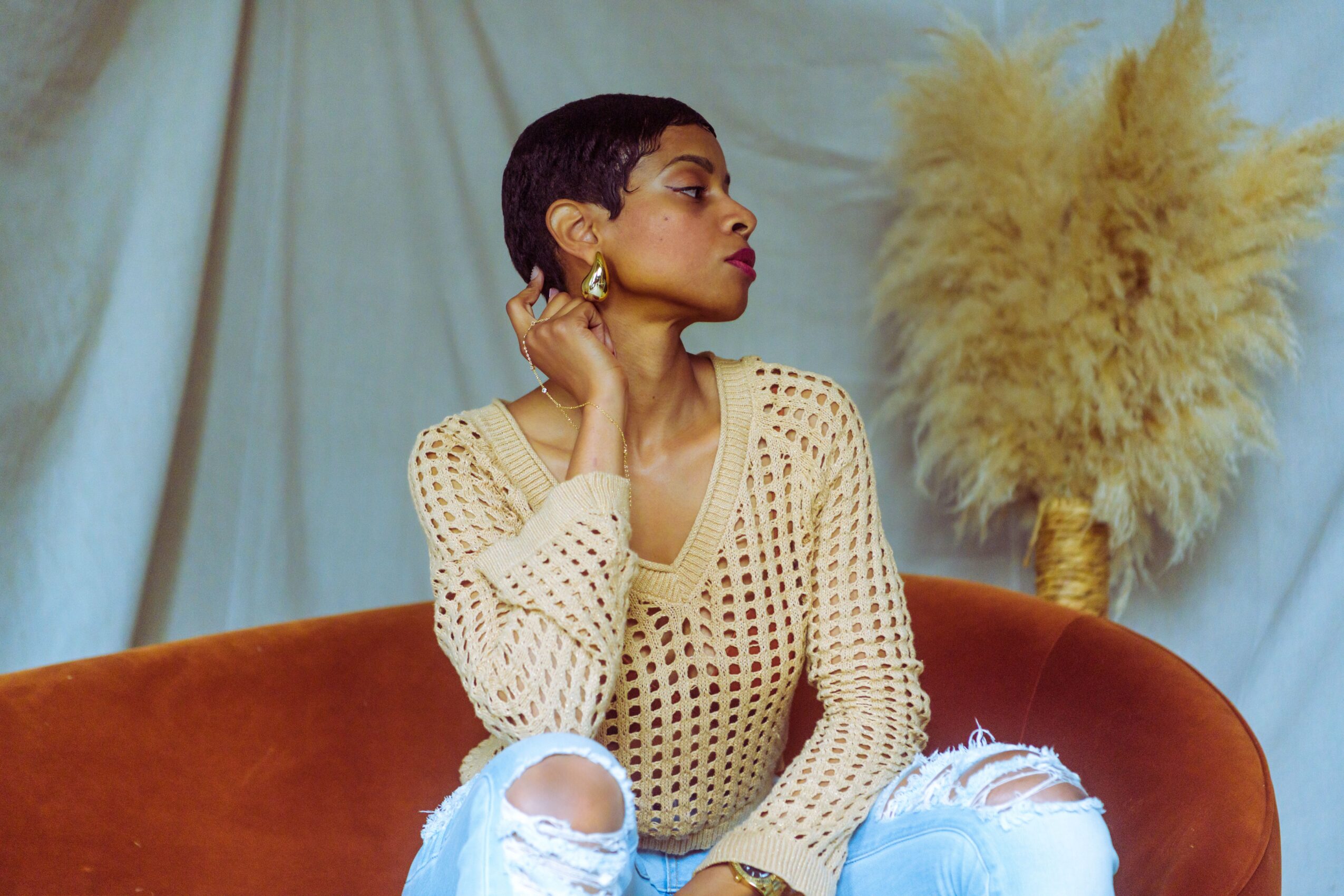 Woman with short hair and earrings sitting on a brown sofa, wearing a cream-colored, perforated sweater, with a large plant in the background.