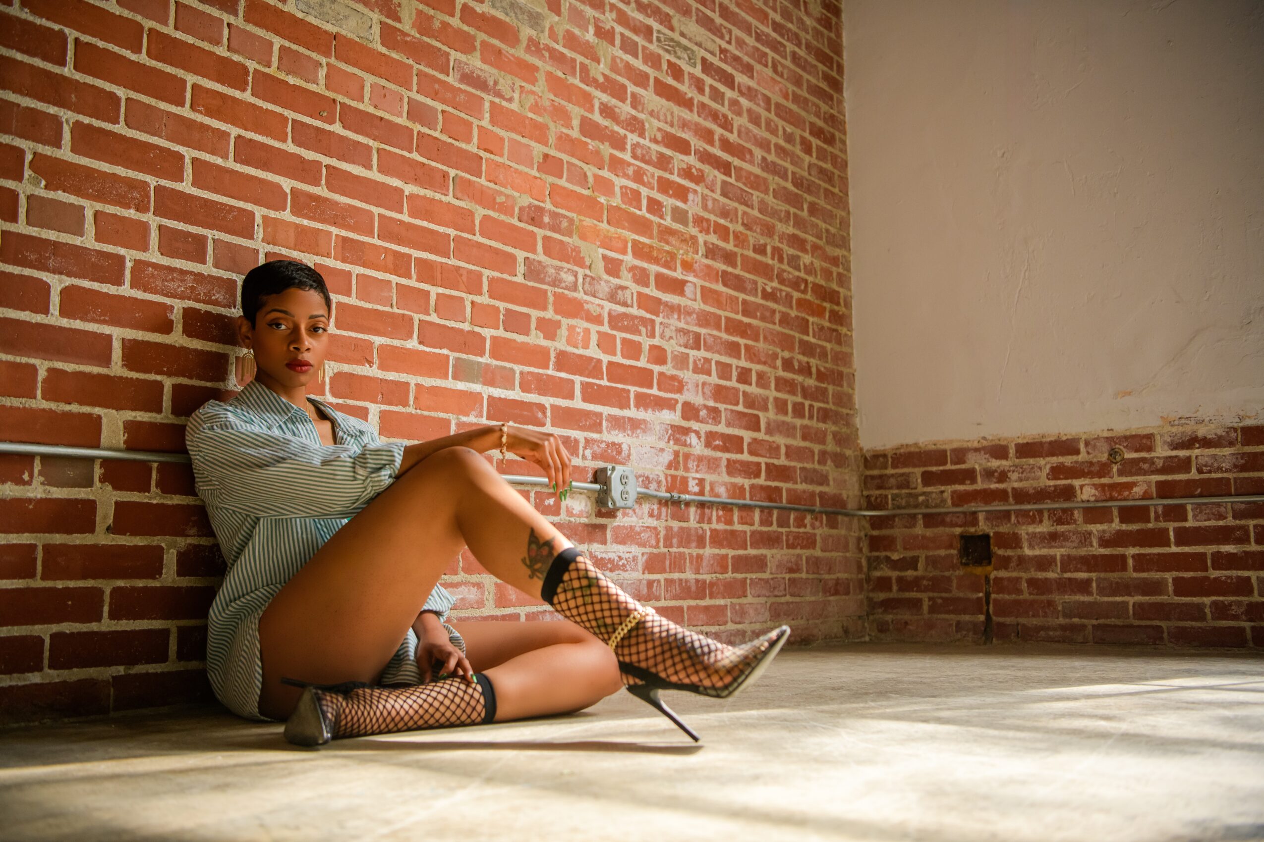 Woman sitting on the floor against a brick wall, holding a drink, with one leg extended and wearing patterned boots.