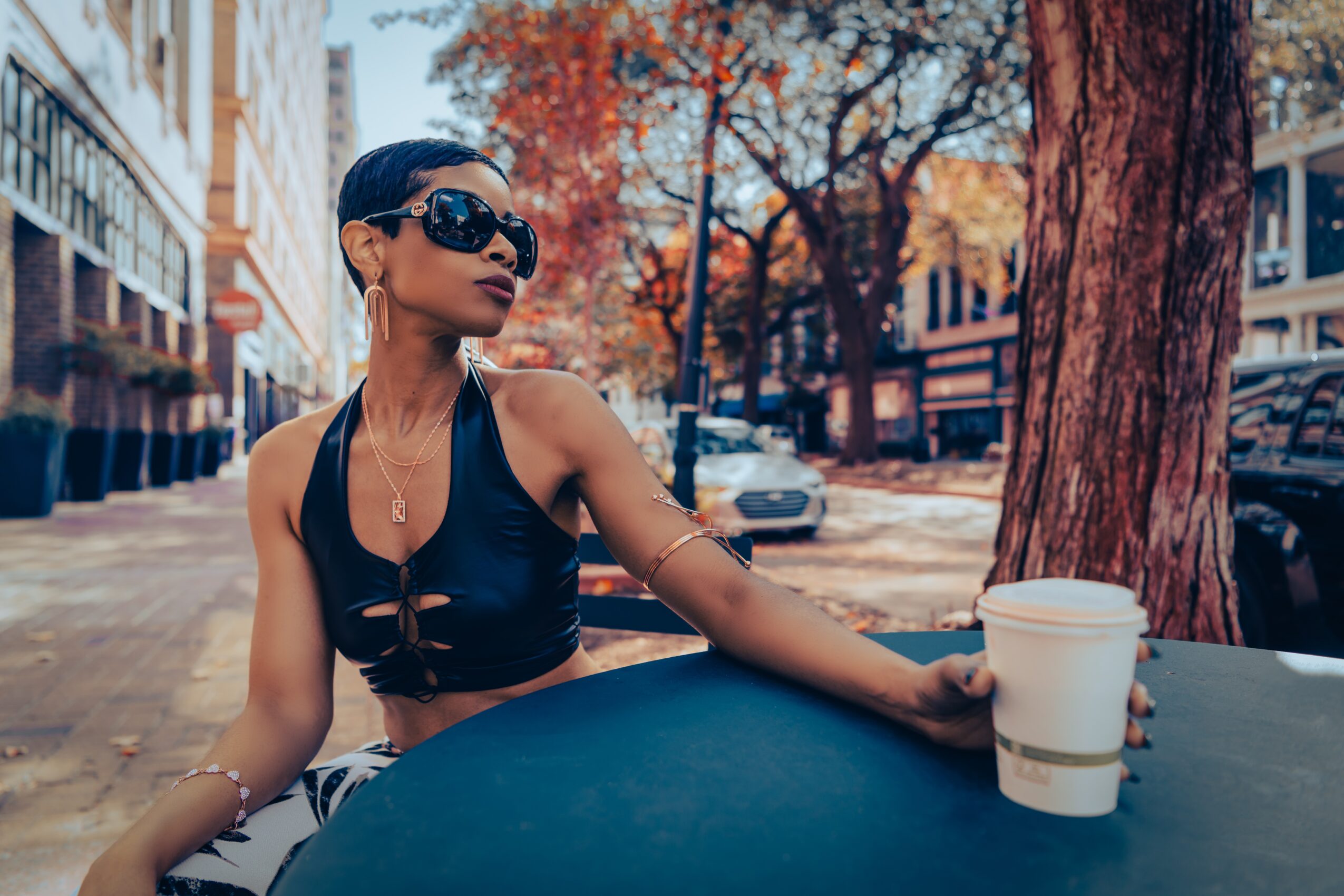 Woman with sunglasses and a black top sitting at a table outdoors, with trees and buildings in the background.