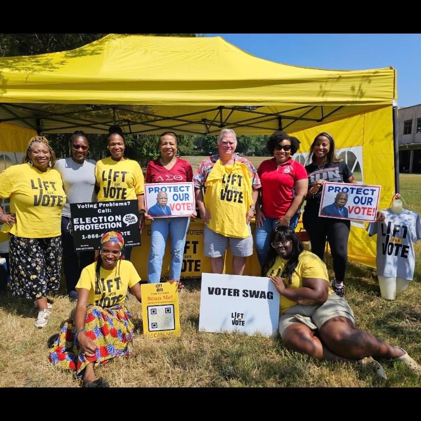 Group of people holding signs promoting voting and election protection outdoors under a yellow canopy, with some sitting and others standing.