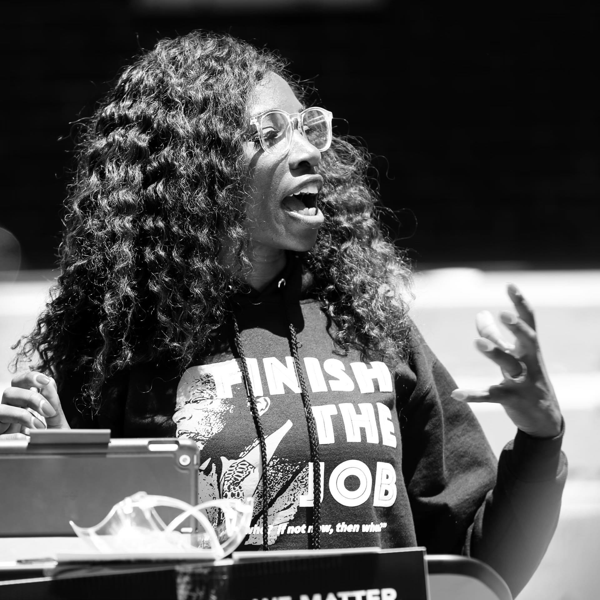 Woman with curly hair and glasses speaking passionately, wearing a hoodie with text, gesturing with hands, in black and white.