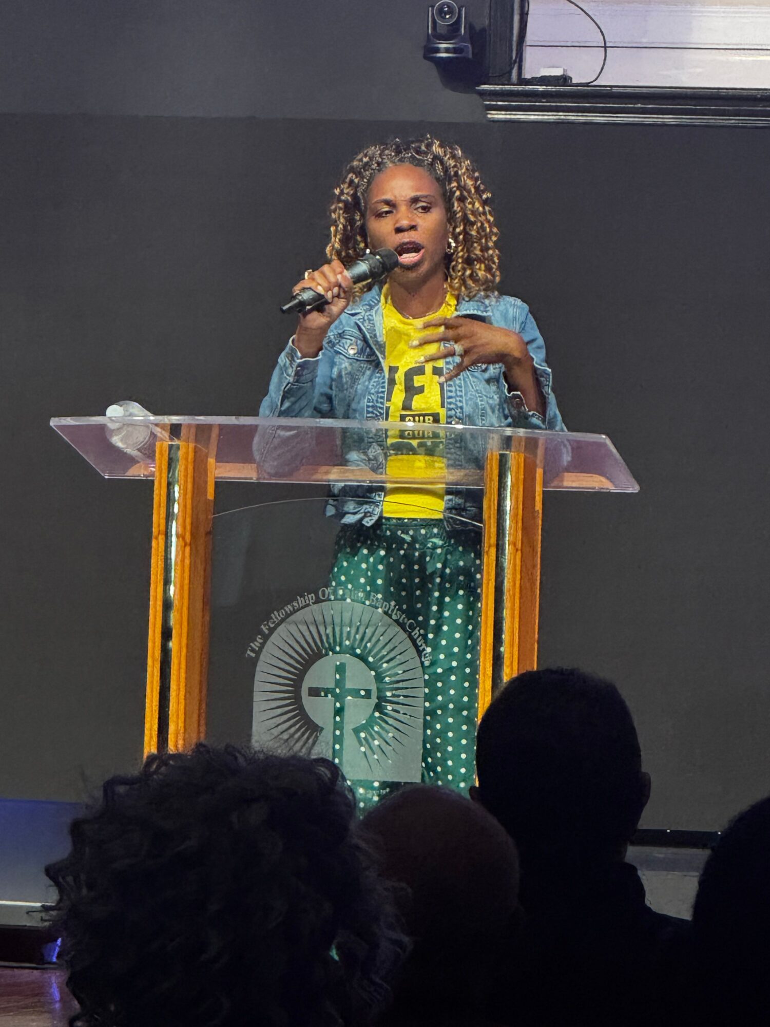 Woman speaking at a podium with a microphone, wearing a yellow shirt and denim jacket, in front of a dark background.